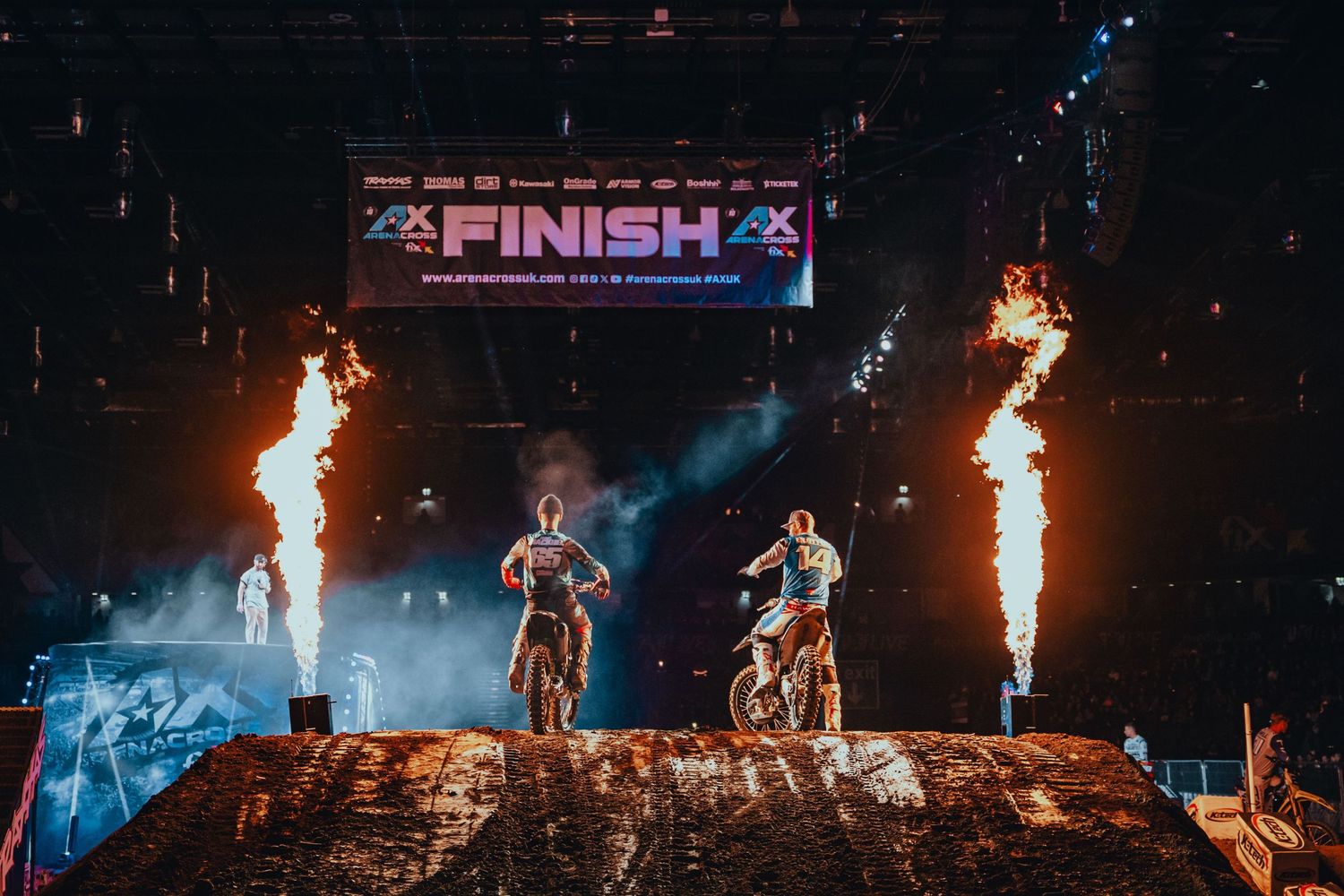 Two motocross riders at a finish line with dramatic fire pyrotechnics at an indoor arena event.