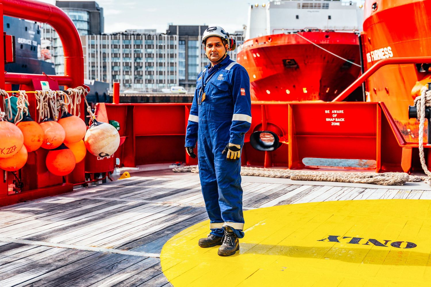 Female crew member in blue coveralls and safety gloves stands on ship helipad at port.