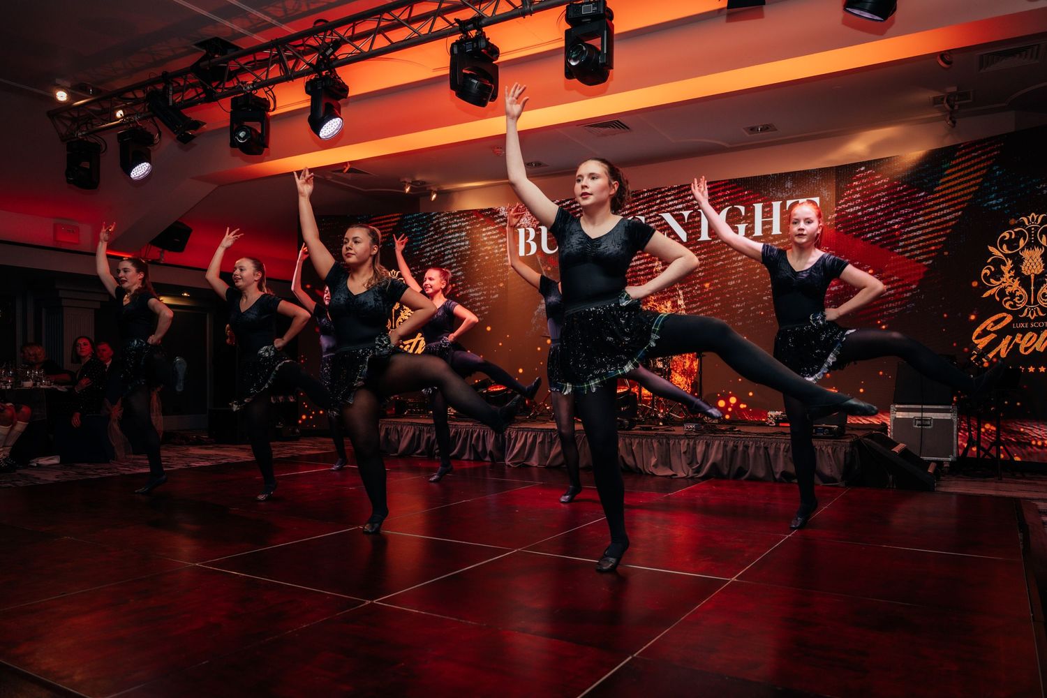 Dancers in black outfits performing on stage at a glamorous evening event with red lighting.