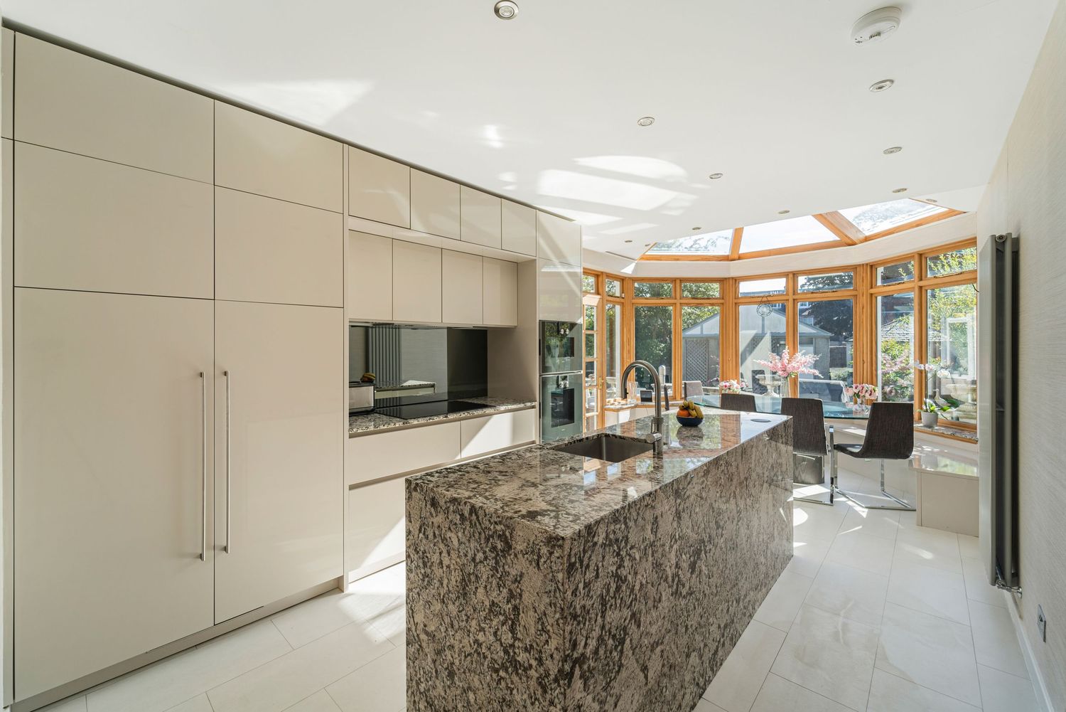 Luxury kitchen with marble island, integrated appliances, cream cabinetry, and conservatory dining area beyond.