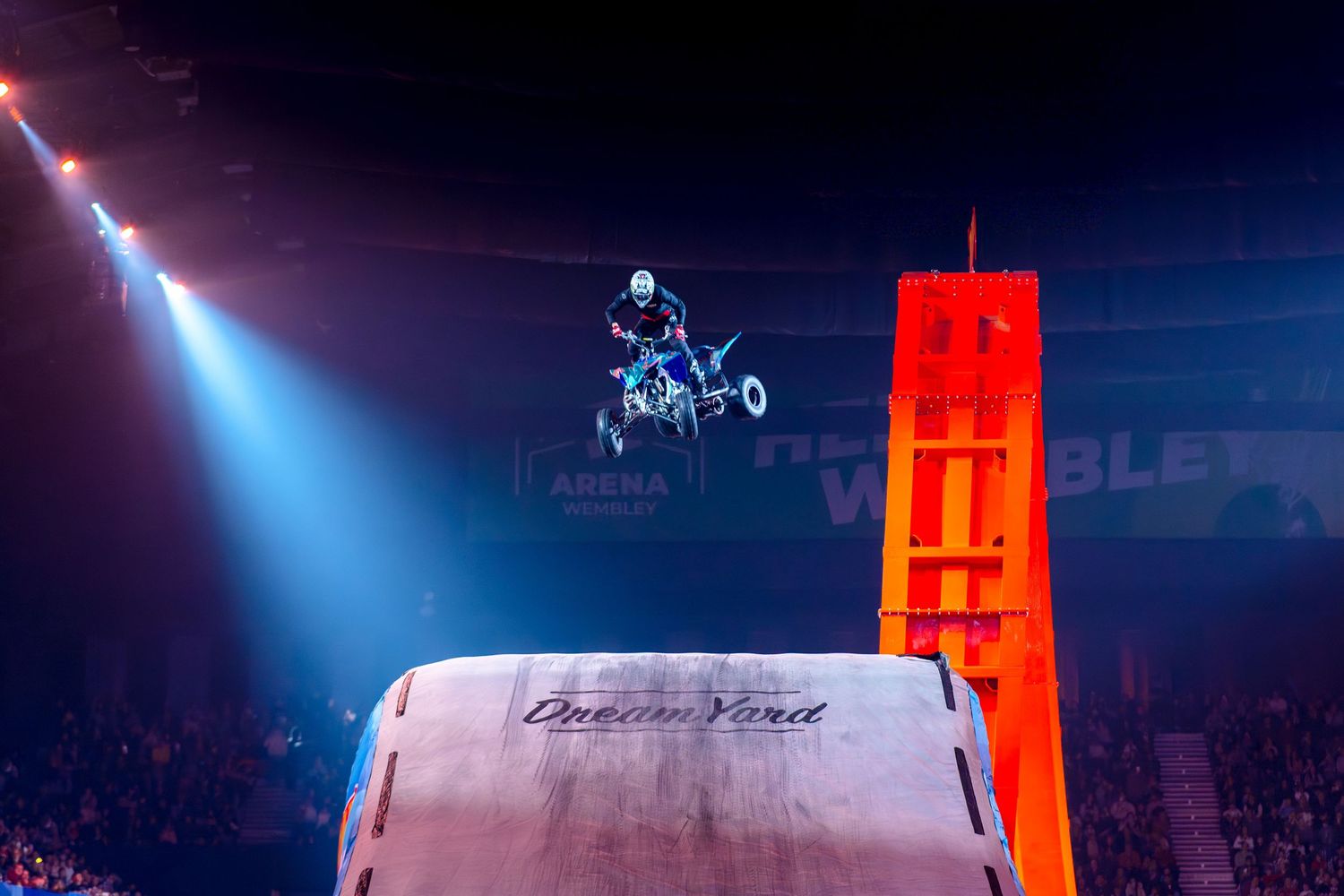 A quad bike rider launches off a Dream Yard ramp in a dramatic indoor arena stunt show.