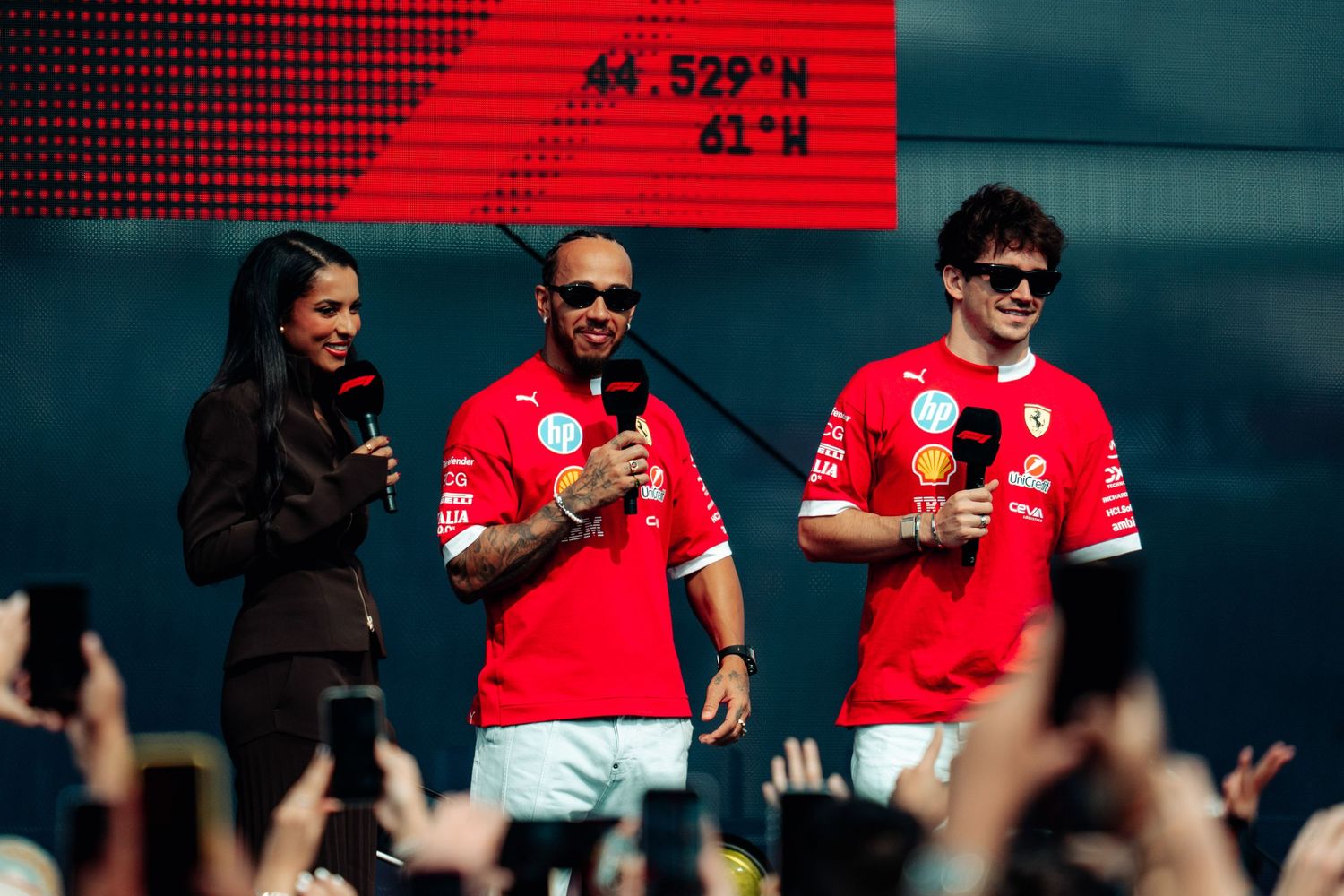 Two Ferrari F1 drivers in red team shirts speak on stage with a female host at a fan event.