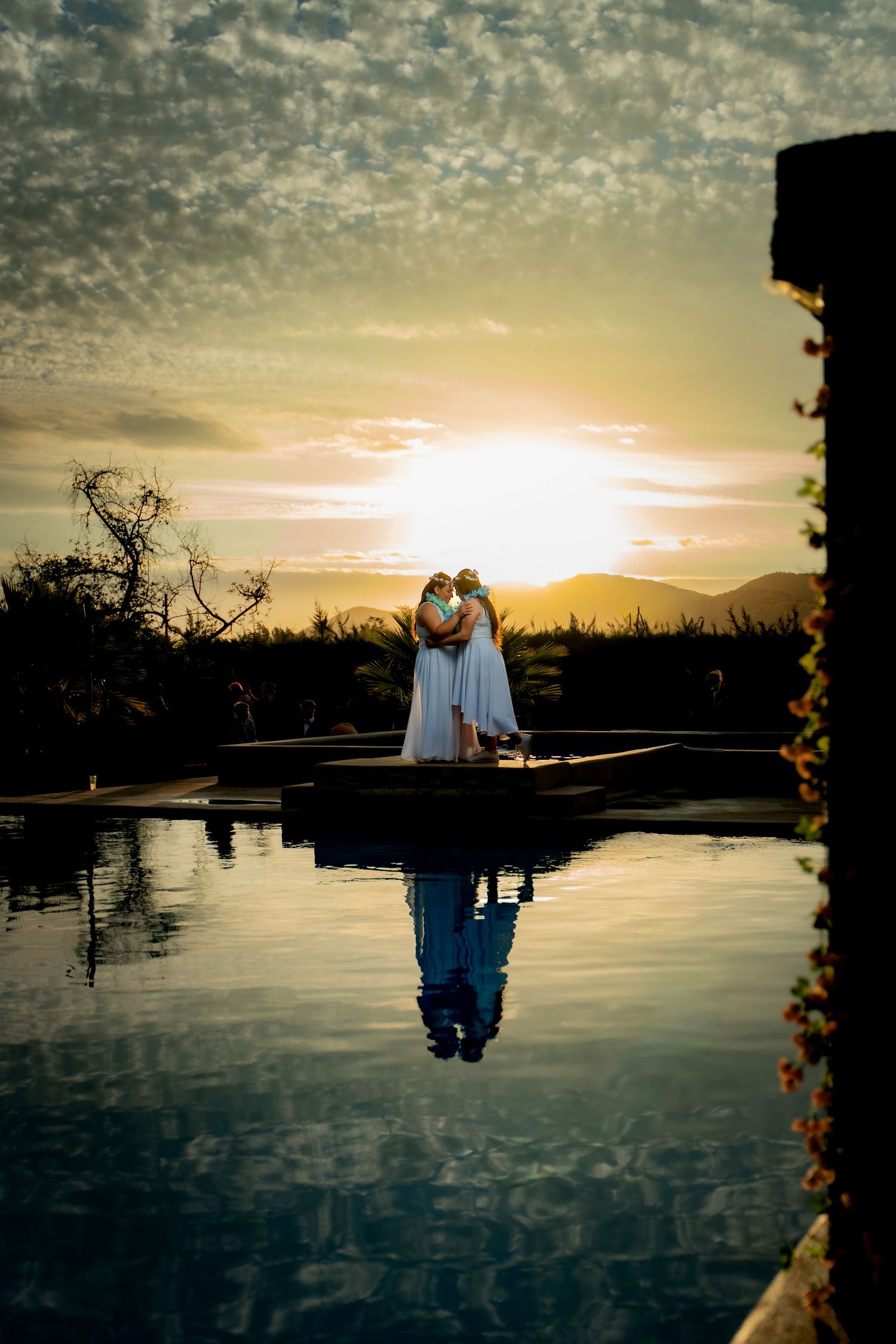 A couple shares an intimate moment at sunset by a reflective pool with dramatic cloudy skies.
