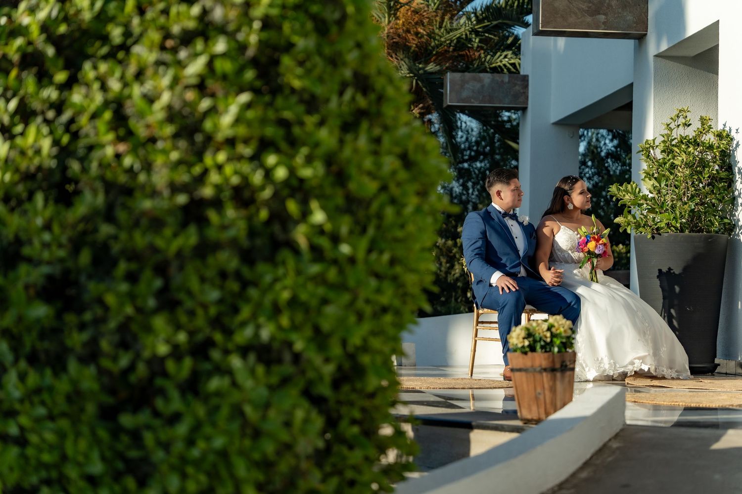 A wedding couple shares a tender moment in front of a white building with lush greenery.