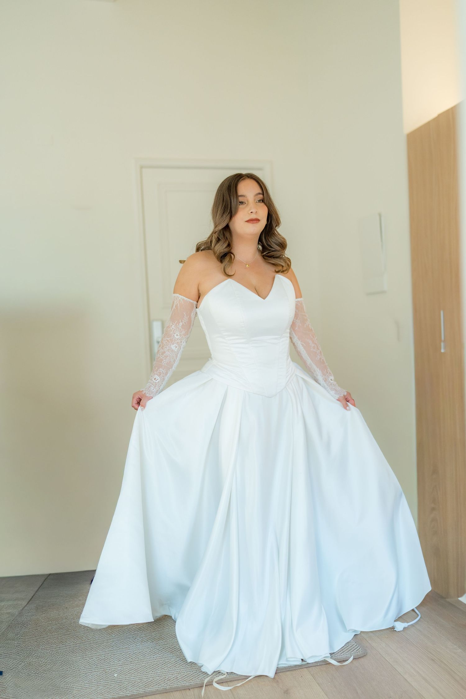 Bride in a white satin A-line gown with lace sleeves posing indoors.