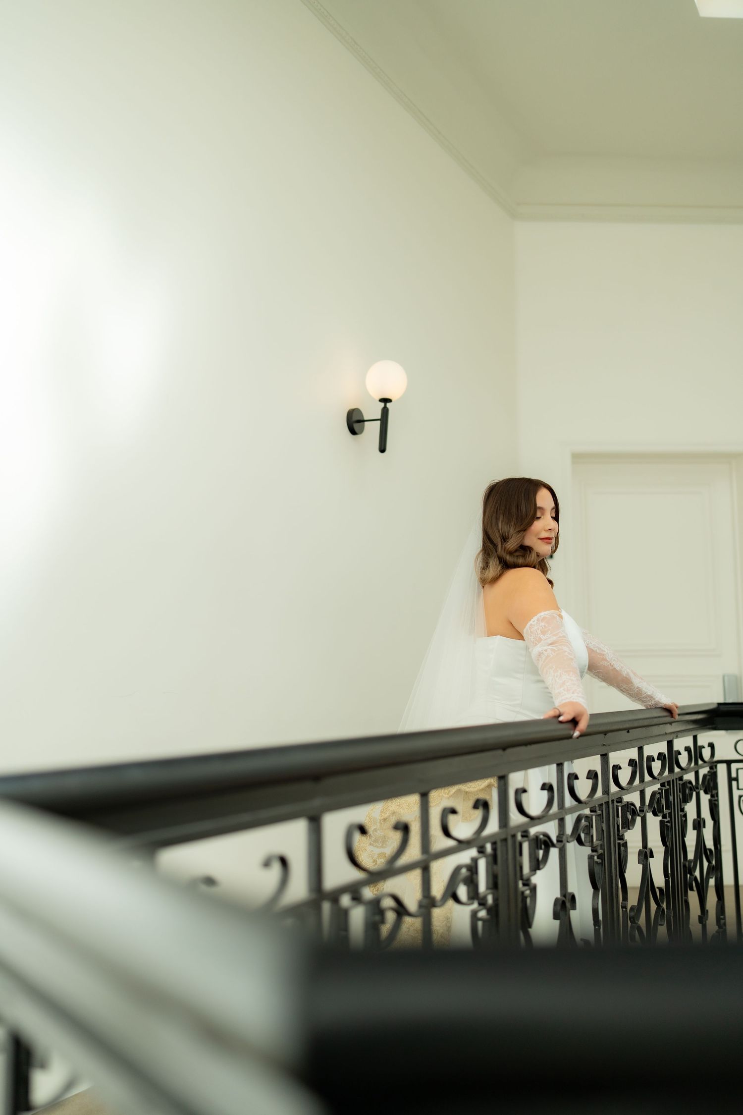 Elegant bride in white off-shoulder gown with lace gloves descending an ornate staircase gracefully.