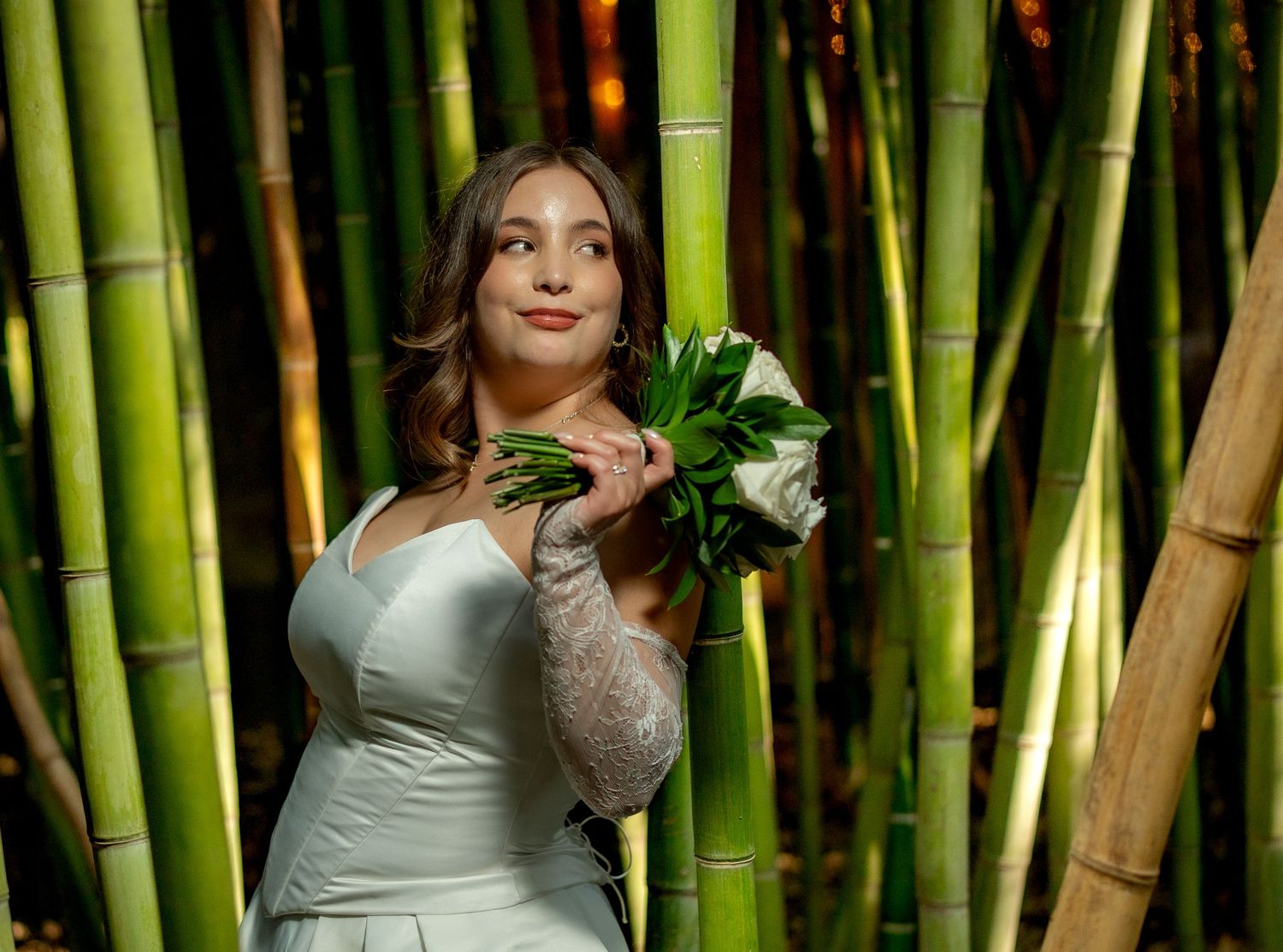 Bride in white gown holding bouquet, posing among tall bamboo trees in outdoor wedding photo.