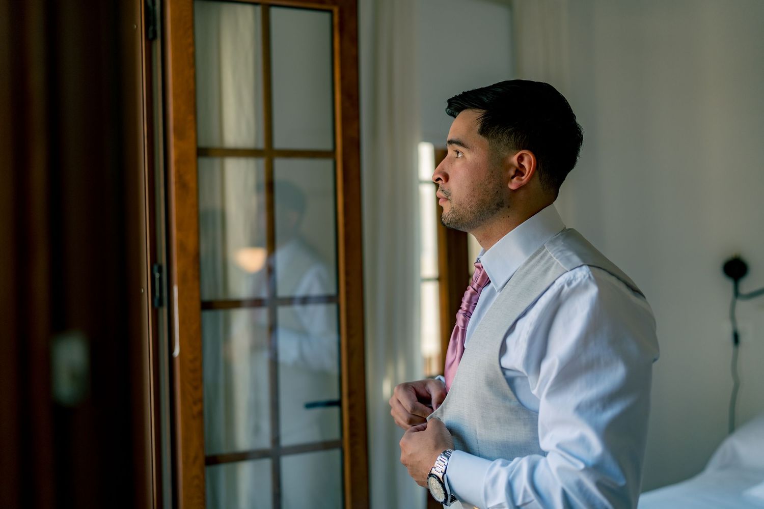 Groom in white dress shirt adjusting his pink tie while gazing out a wooden framed window.