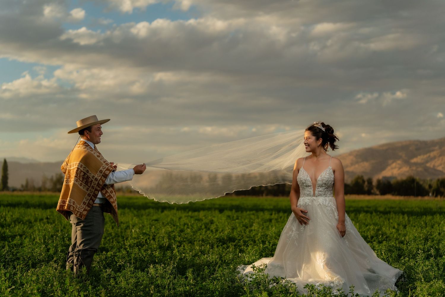 Couple in wedding attire poses in a lush green field under dramatic cloudy skies at sunset.