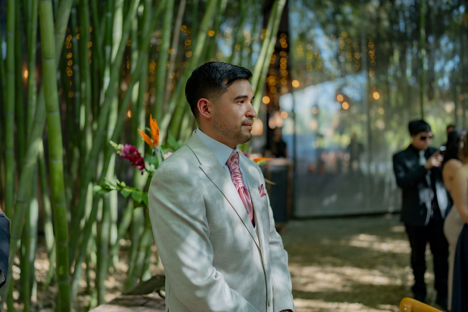 Handsome groom in light gray suit watching bride walk down aisle at outdoor bamboo wedding.