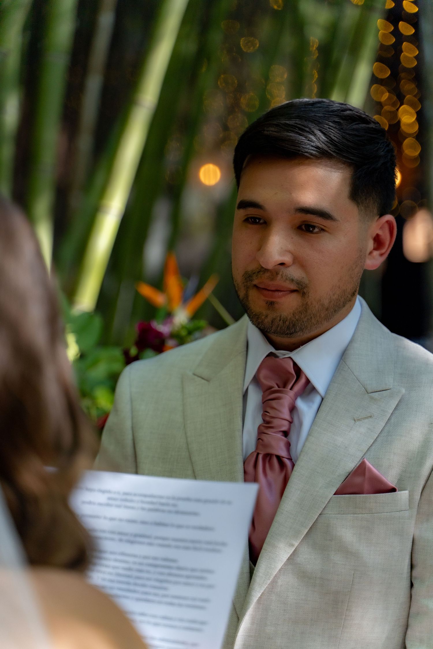 Groom in beige suit with pink tie listens attentively during outdoor bamboo forest ceremony.