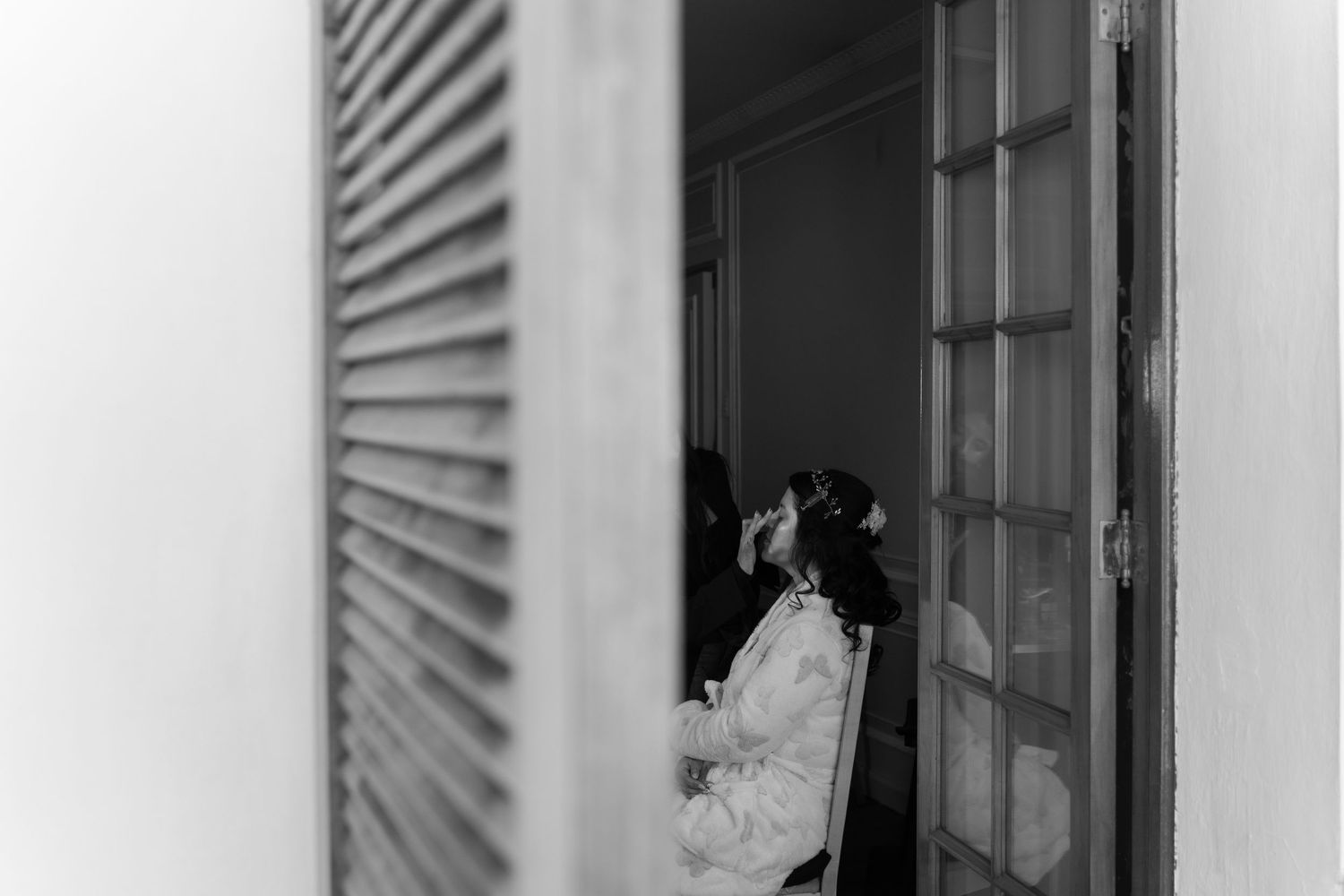 Black and white photograph of a person standing in a doorway with window shutters casting dramatic shadows.