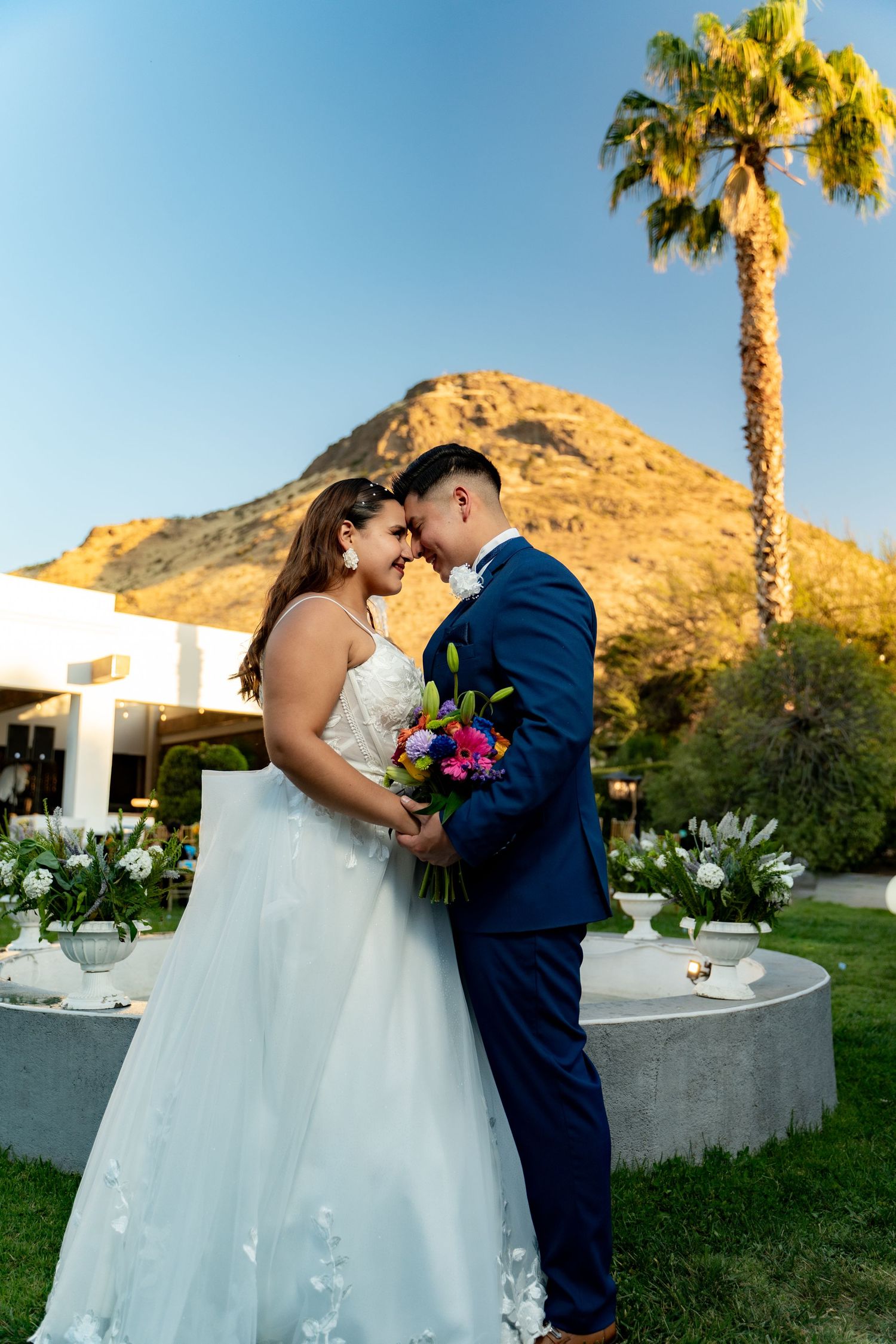 Newlyweds share intimate moments in a garden setting with palm trees and mountain views.