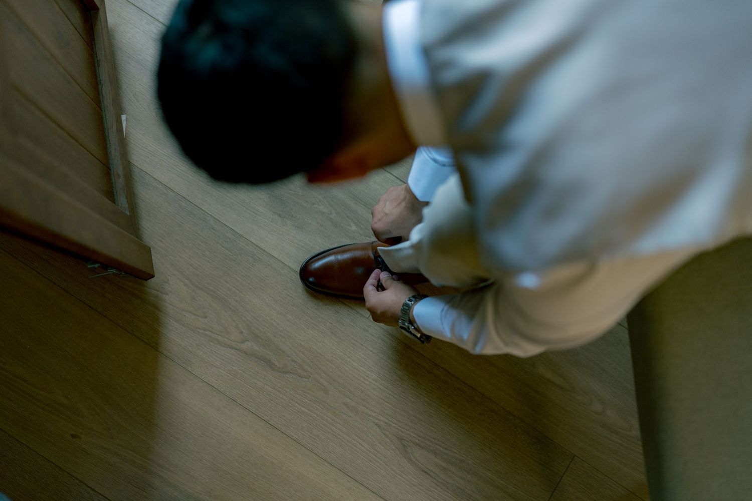 Groom bending down to put on brown leather dress shoes on a light wood floor before wedding.