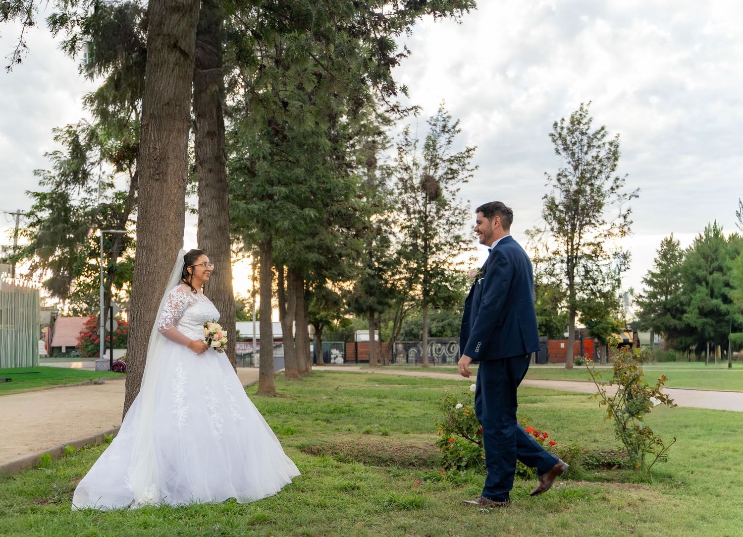 A couple in wedding attire shares a moment in a park with tall pine trees.