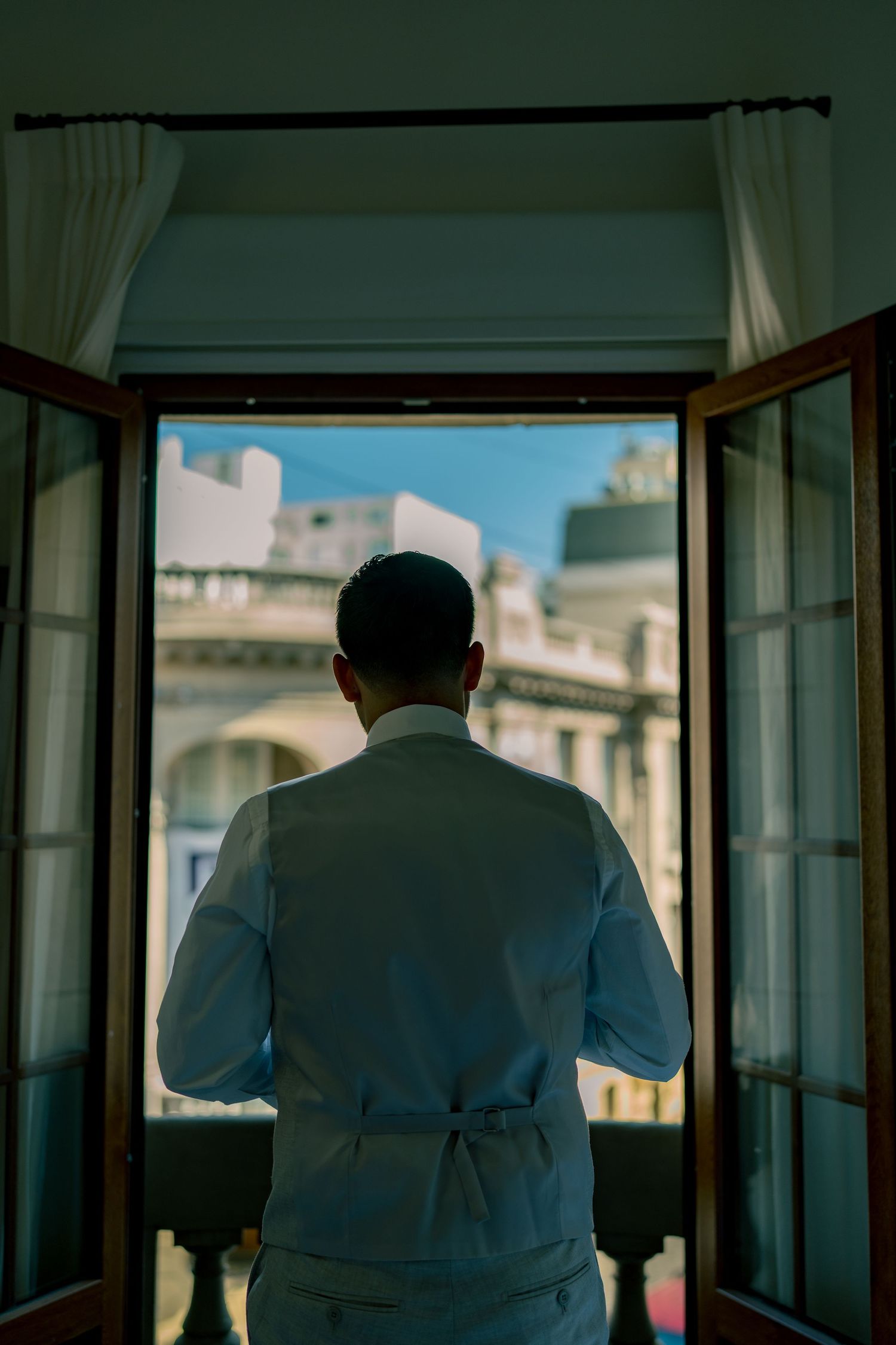 Groom in light gray suit stands at open balcony doors overlooking elegant architecture on his wedding day.