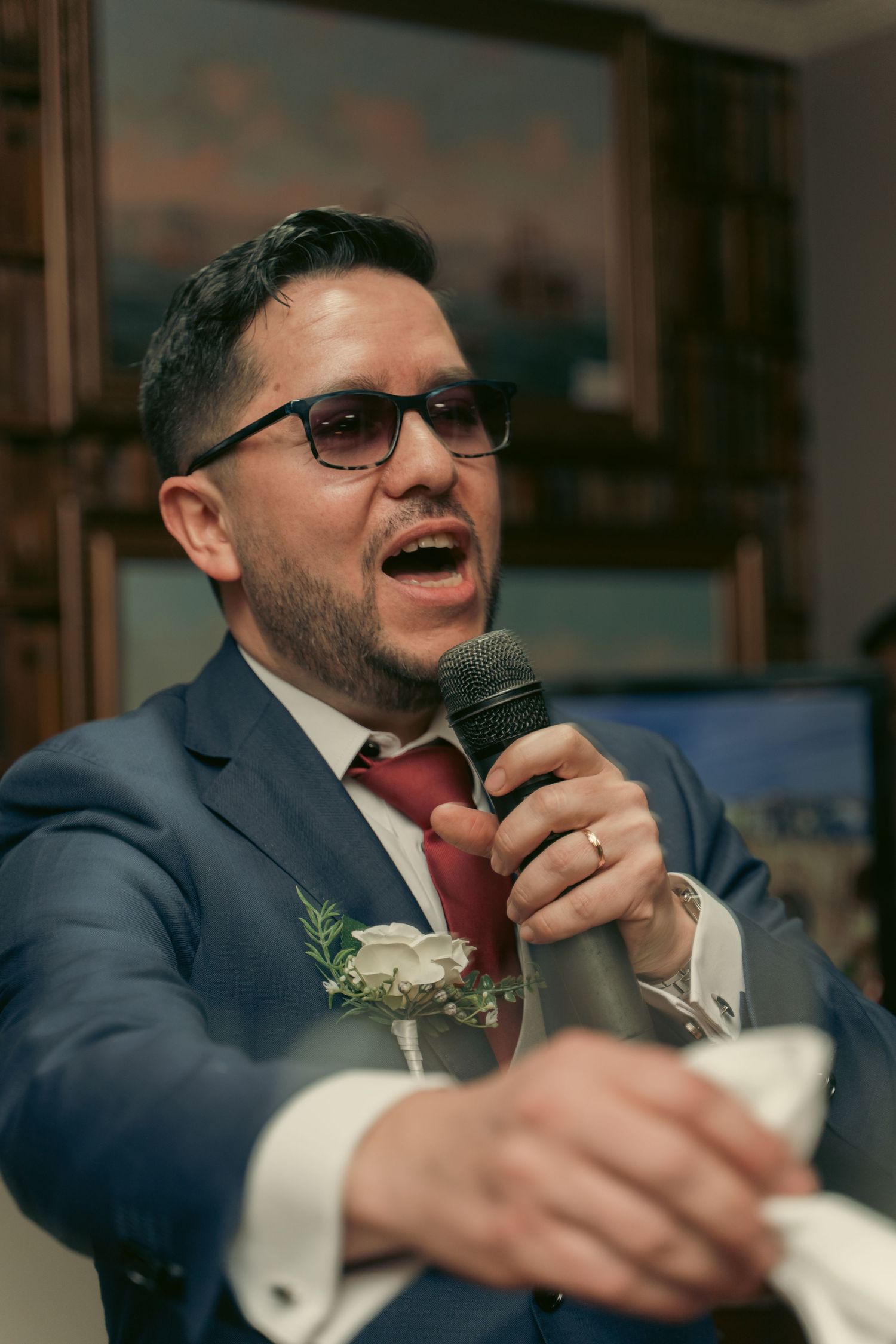 Groom in navy suit and red tie speaks into microphone during wedding reception.