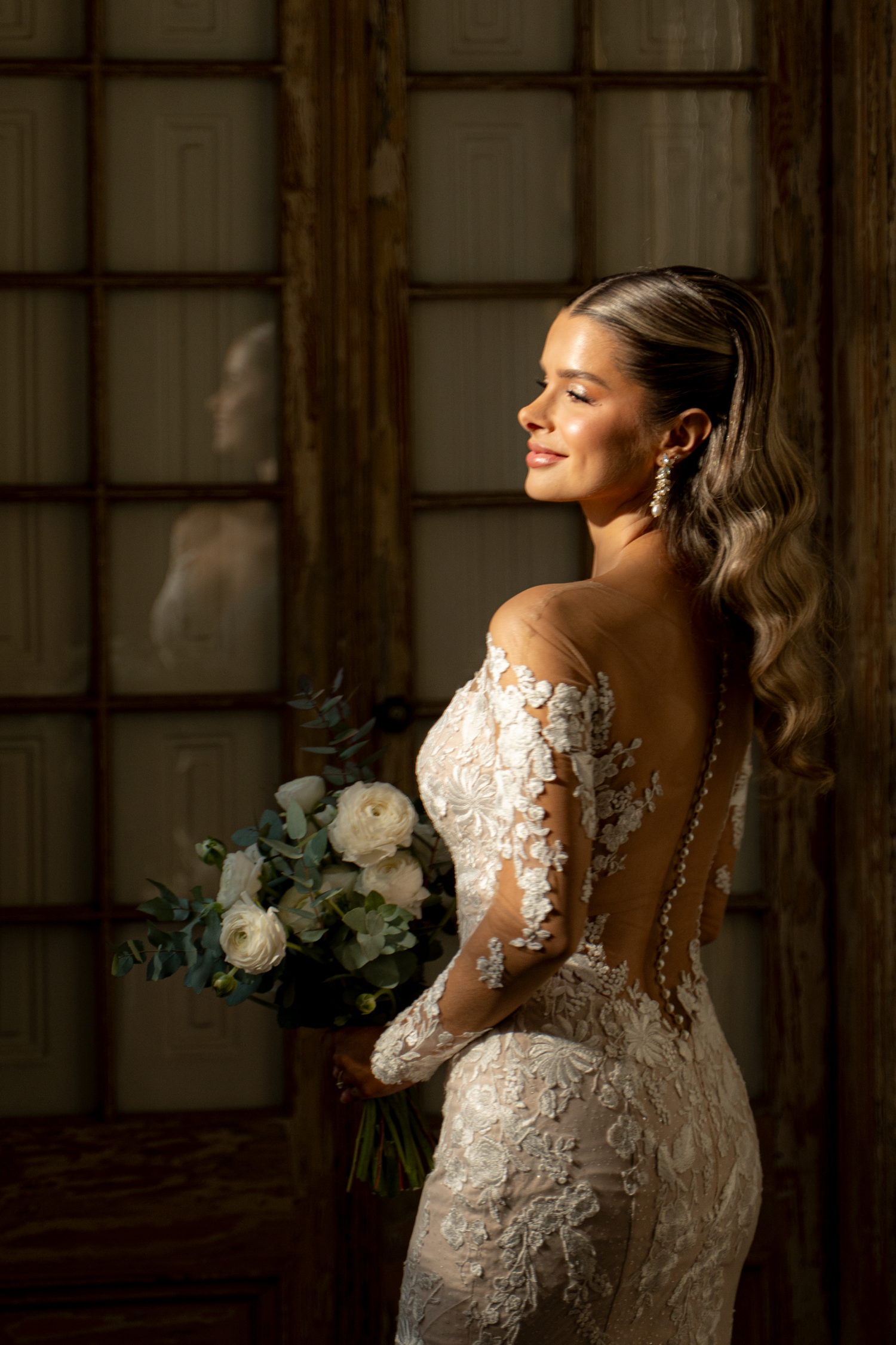 Bride in lace gown poses by window with natural light streaming in.