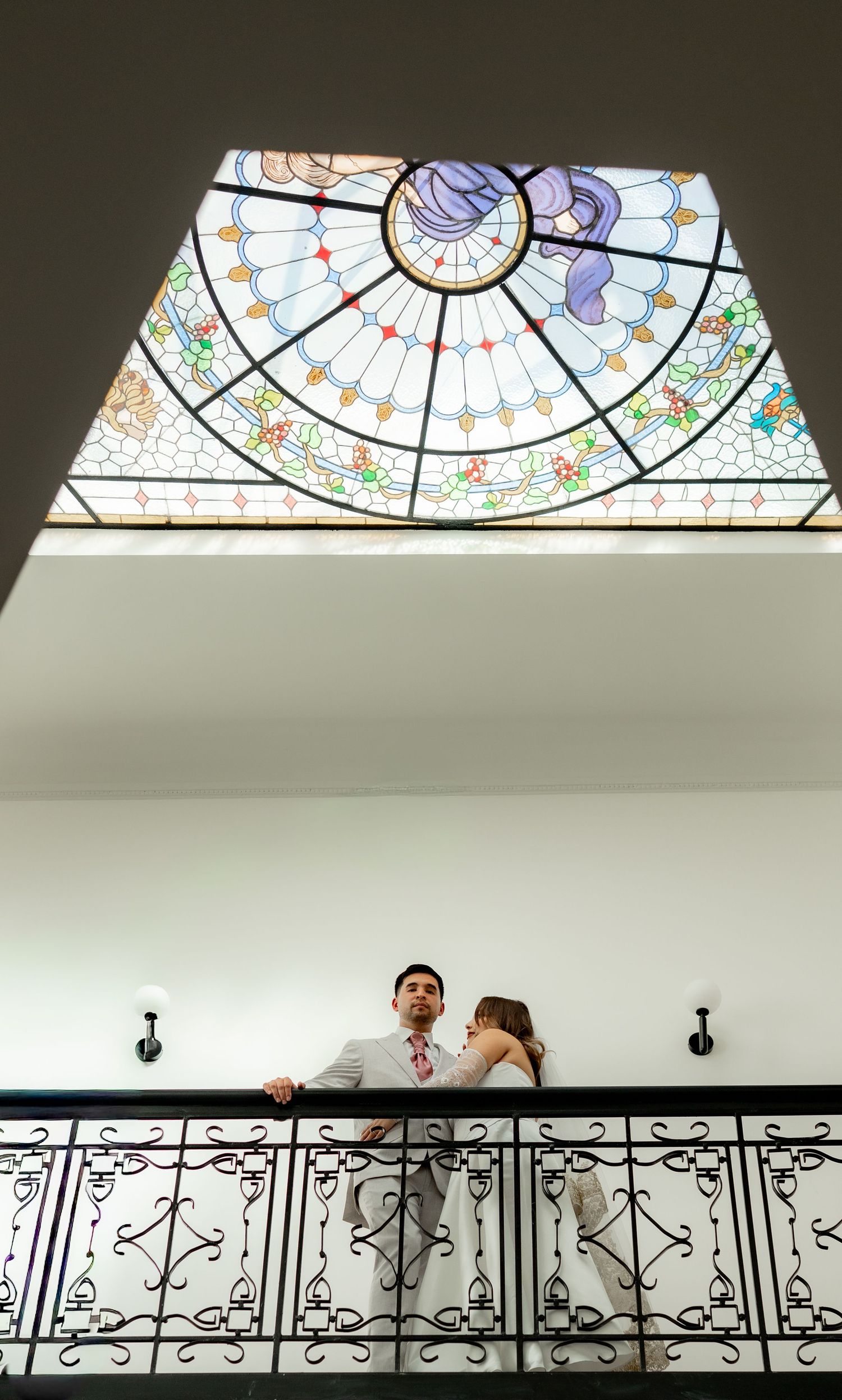 Wedding couple sharing a romantic moment on a balcony beneath a stunning stained glass dome ceiling.