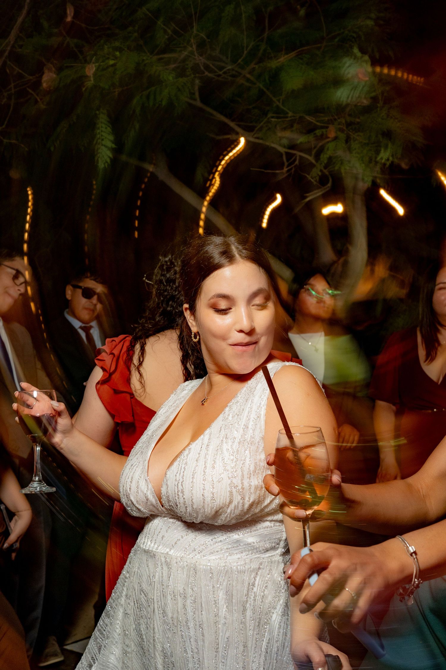 Bride in white gown dancing with champagne glass at outdoor wedding reception under string lights.