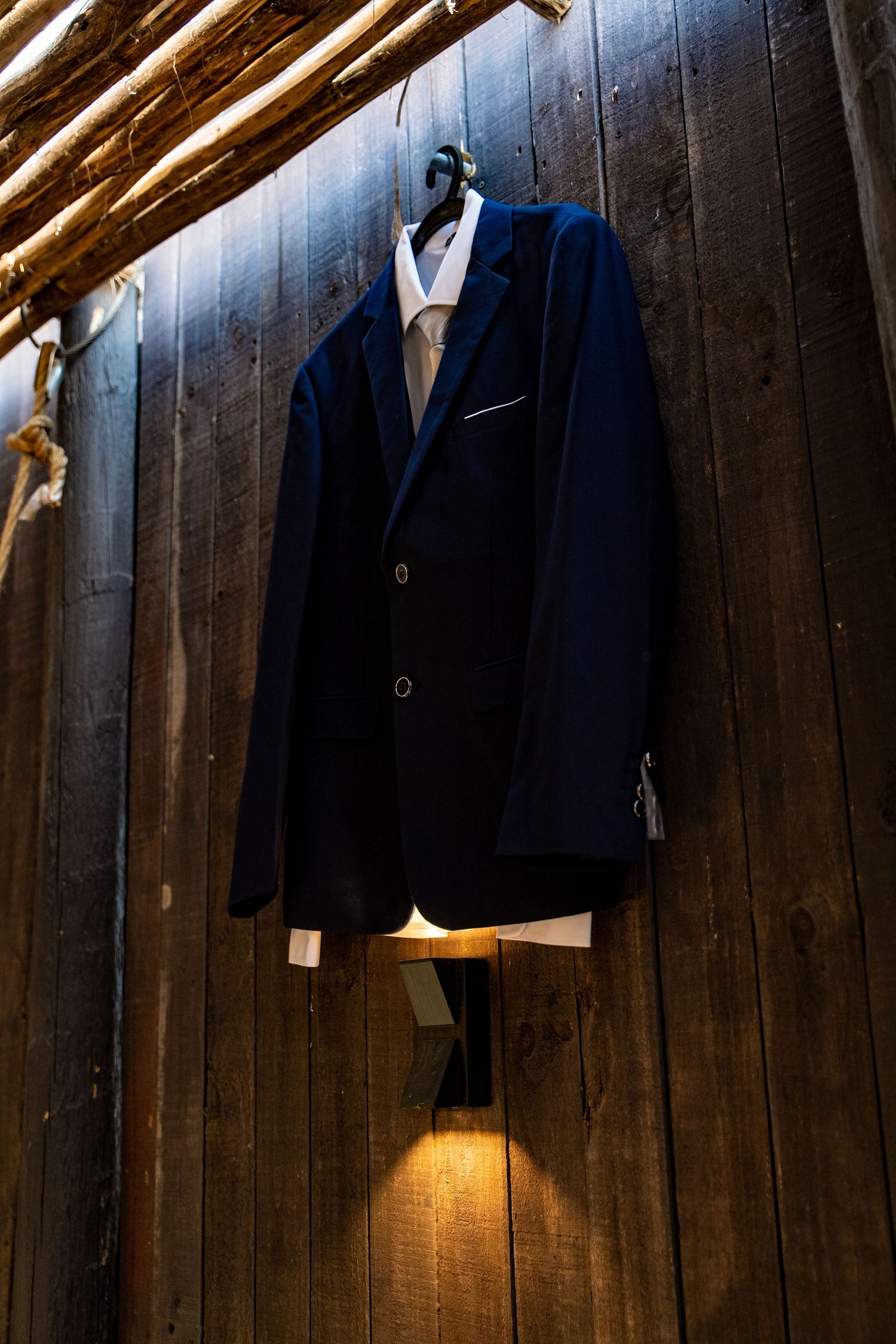 Navy blue wedding suit jacket hanging against wooden wall with dramatic lighting.