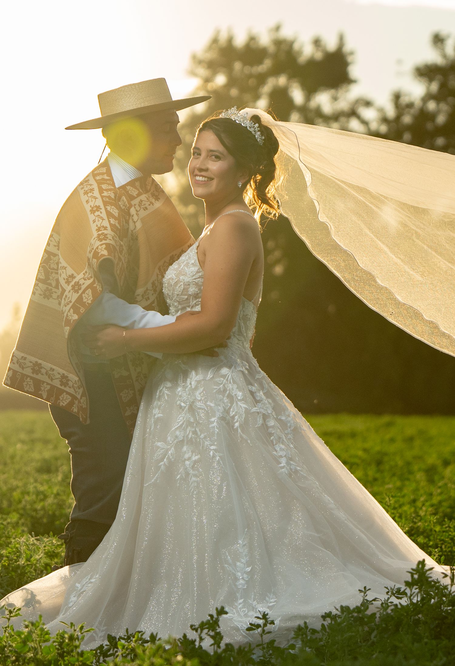 A romantic wedding couple dances together in a sunlit field with flowing white dress and cowboy hat at golden hour.