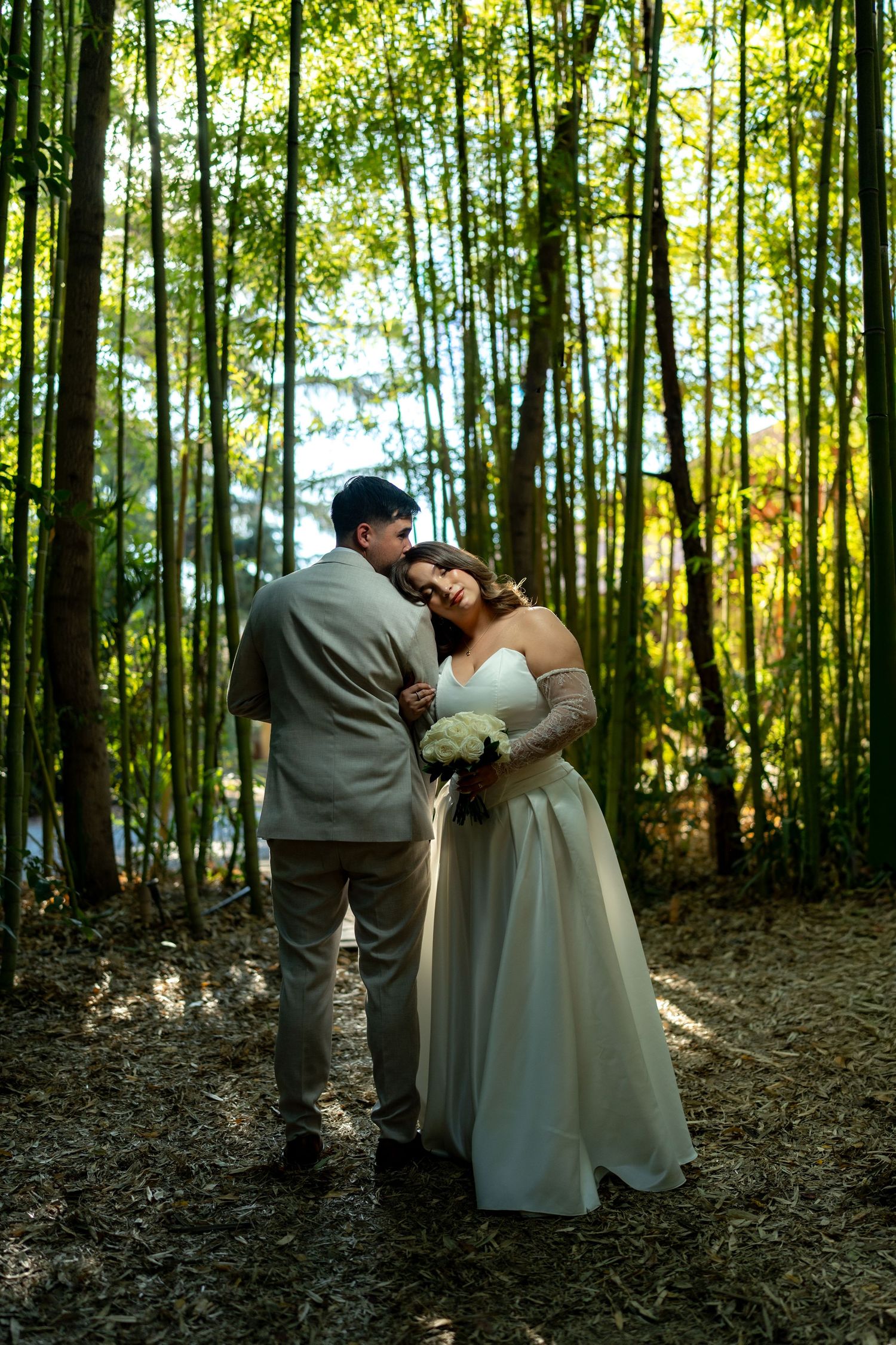 Romantic newlyweds embracing tenderly among tall bamboo trees in sunlit forest after wedding.
