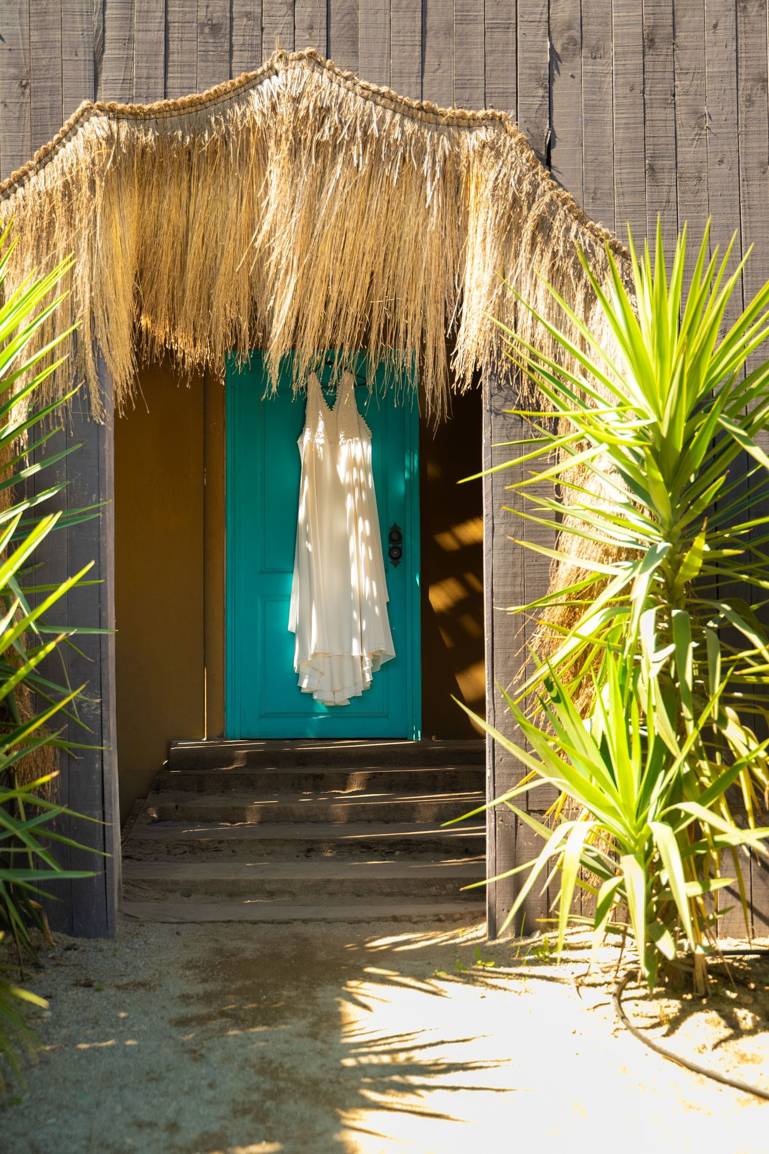 White wedding dress hanging in a tropical doorway with thatched roof and palm plants.