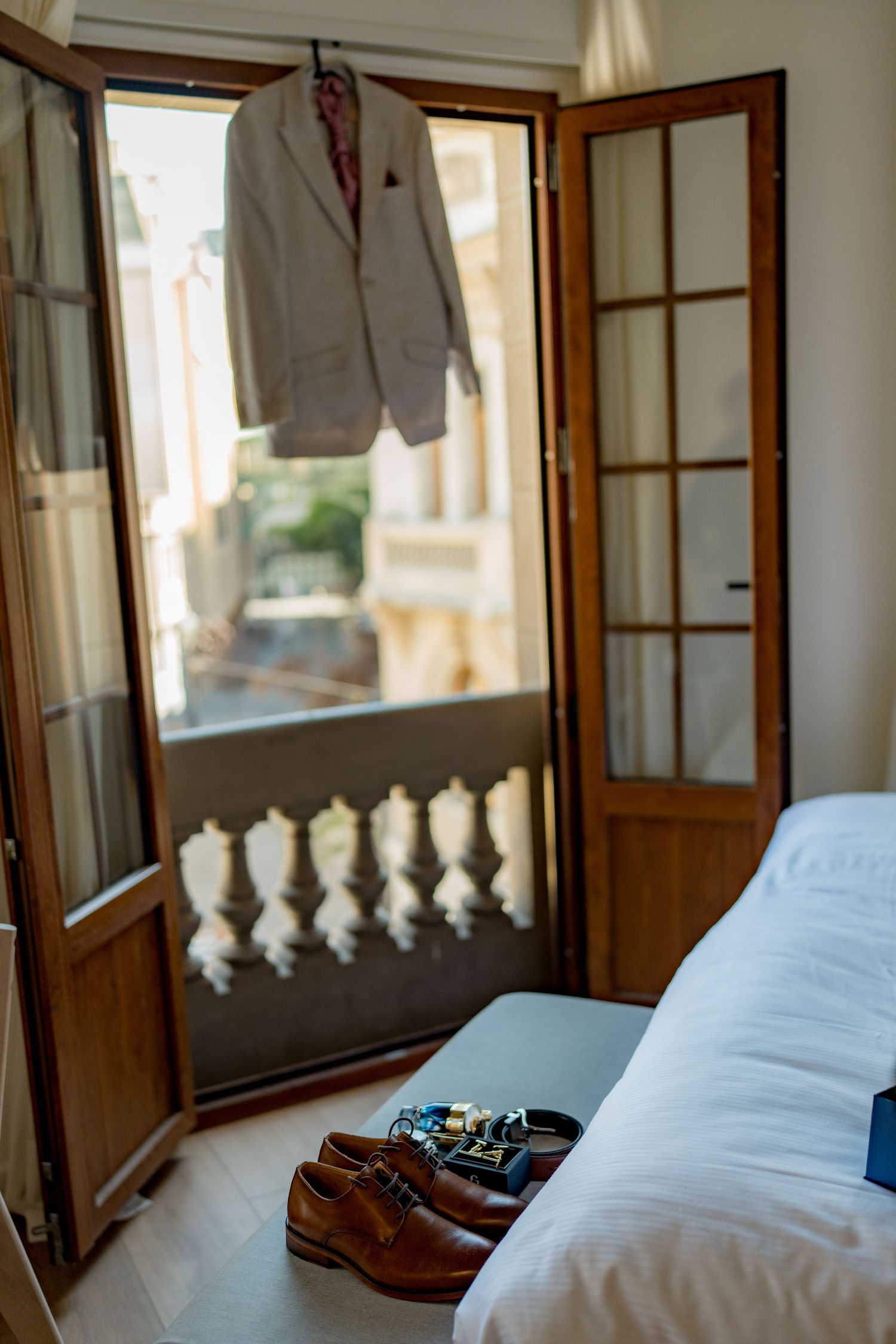 Groom's wedding suit hanging on balcony door with brown leather dress shoes and accessories on white bed.