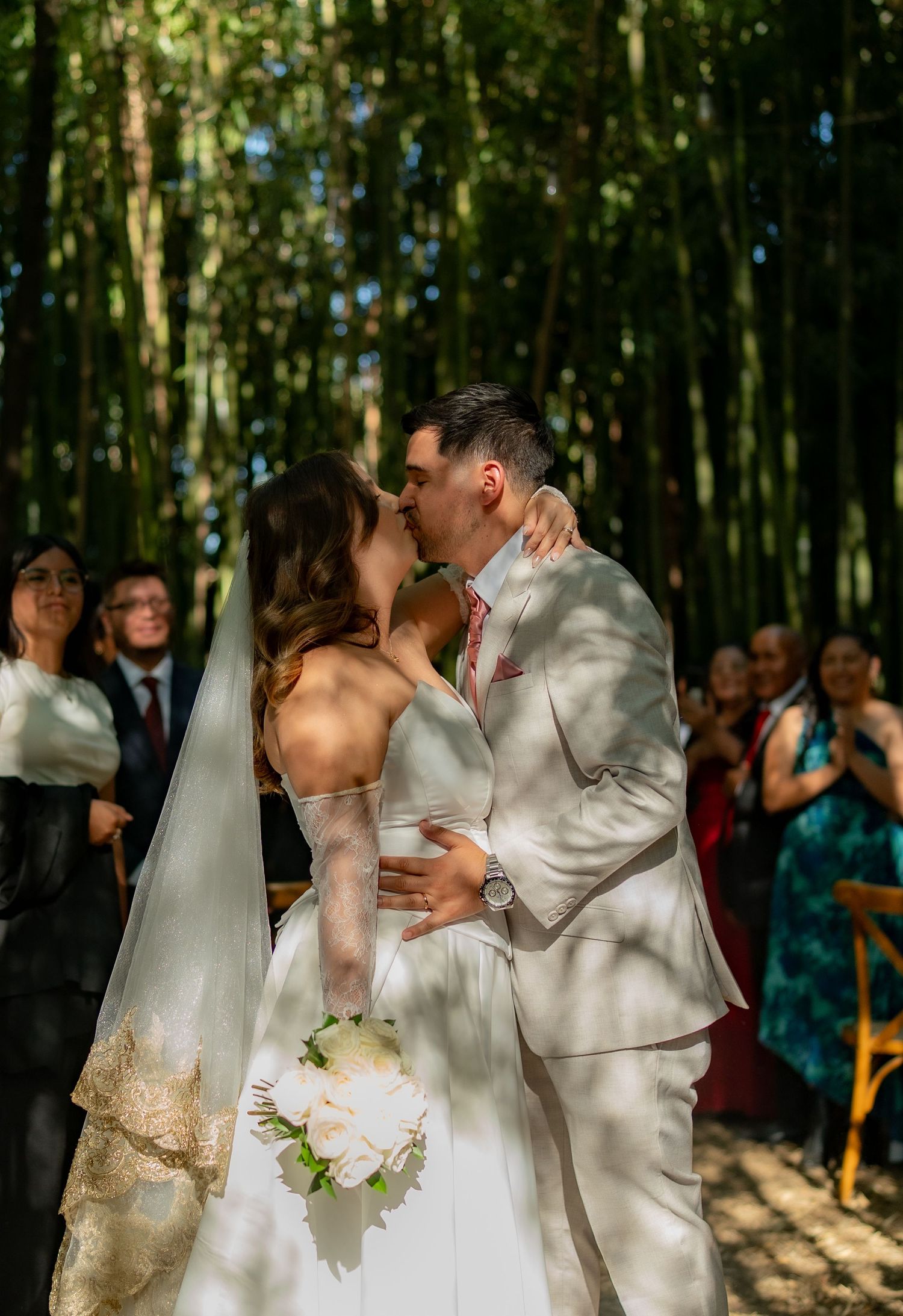 Bride and groom kissing during outdoor bamboo forest wedding ceremony while guests applaud in background.