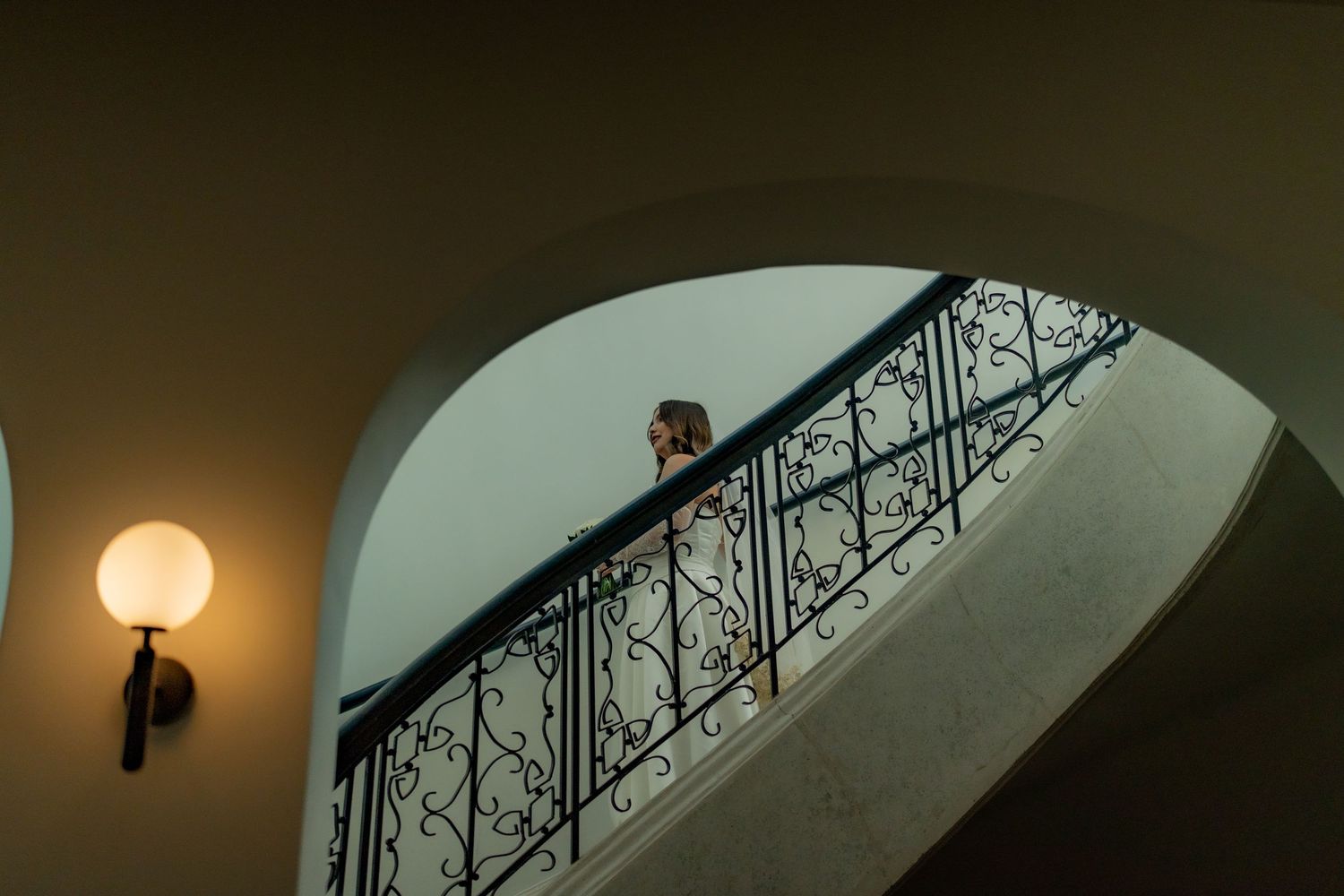 Bride descends ornate wrought iron staircase in a grand venue on her wedding day.
