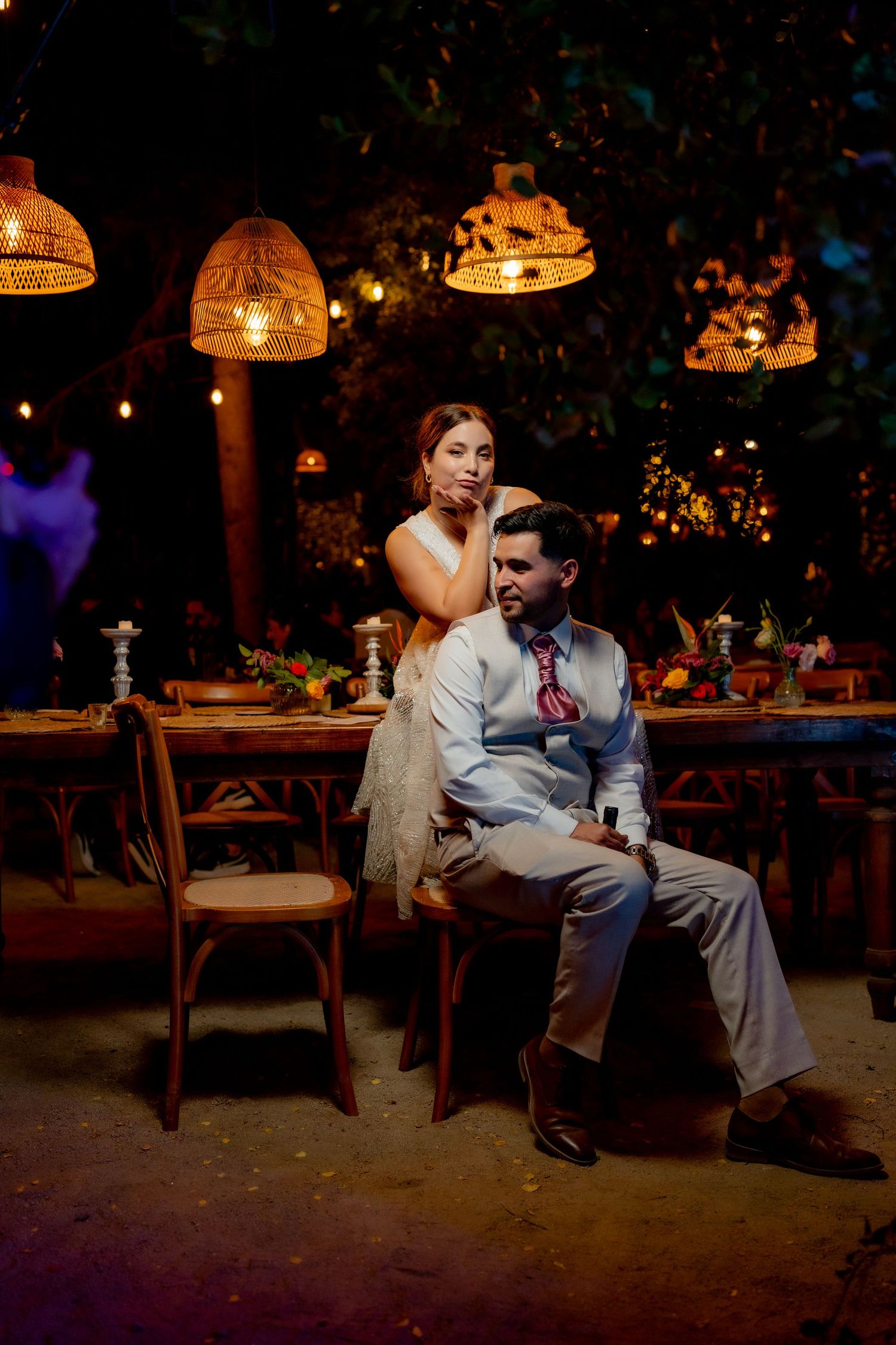 Bride and groom posing elegantly at decorated wedding table under warm wicker pendant lights at night.