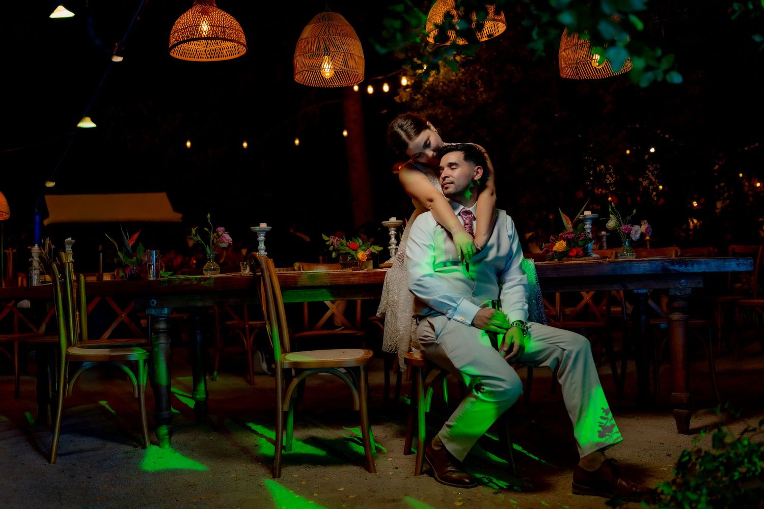Romantic couple embracing on a chair in a dimly lit outdoor restaurant with green lights at night.