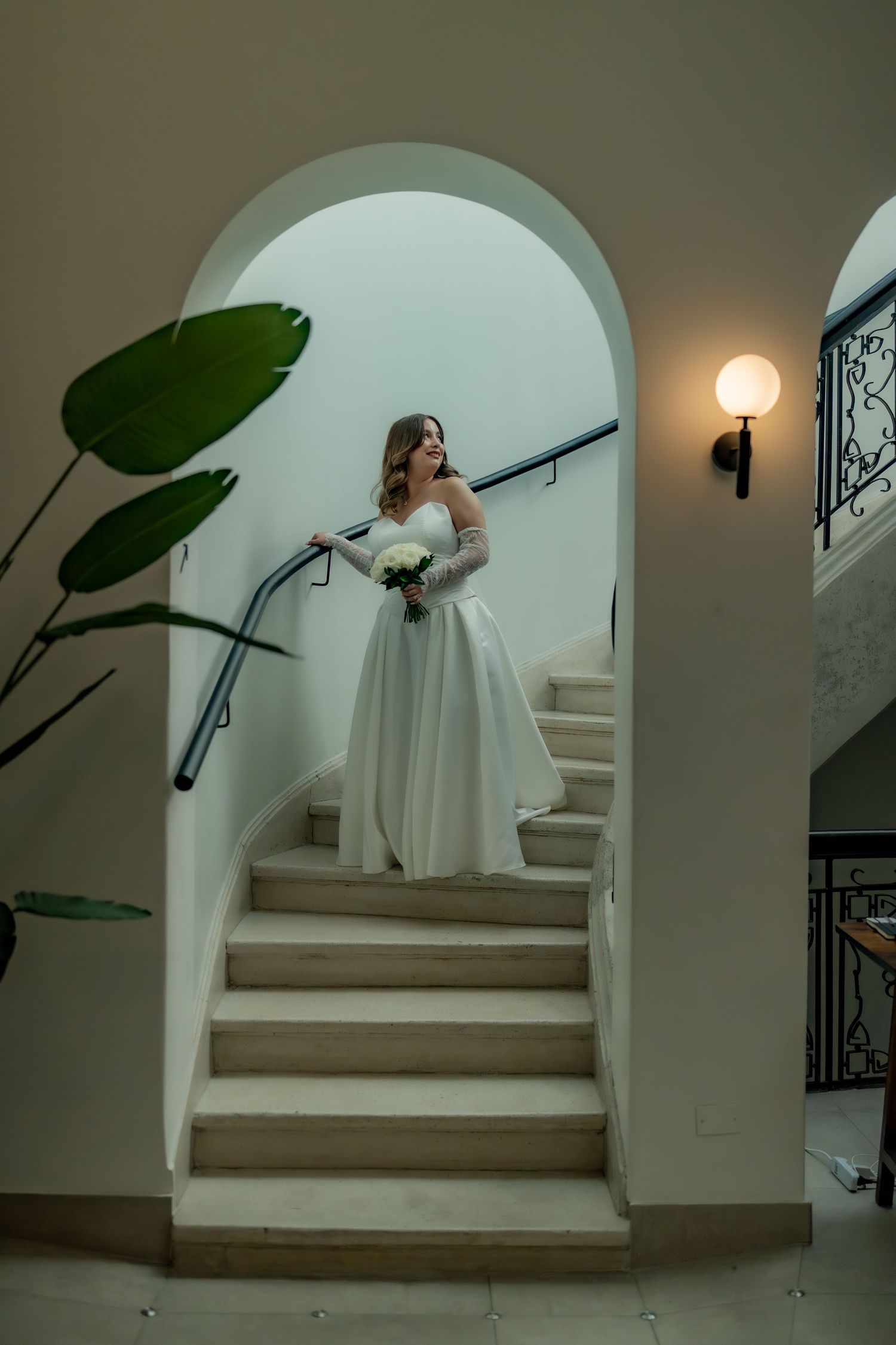Bride in white ball gown with lace sleeves poses on grand staircase holding white bouquet.