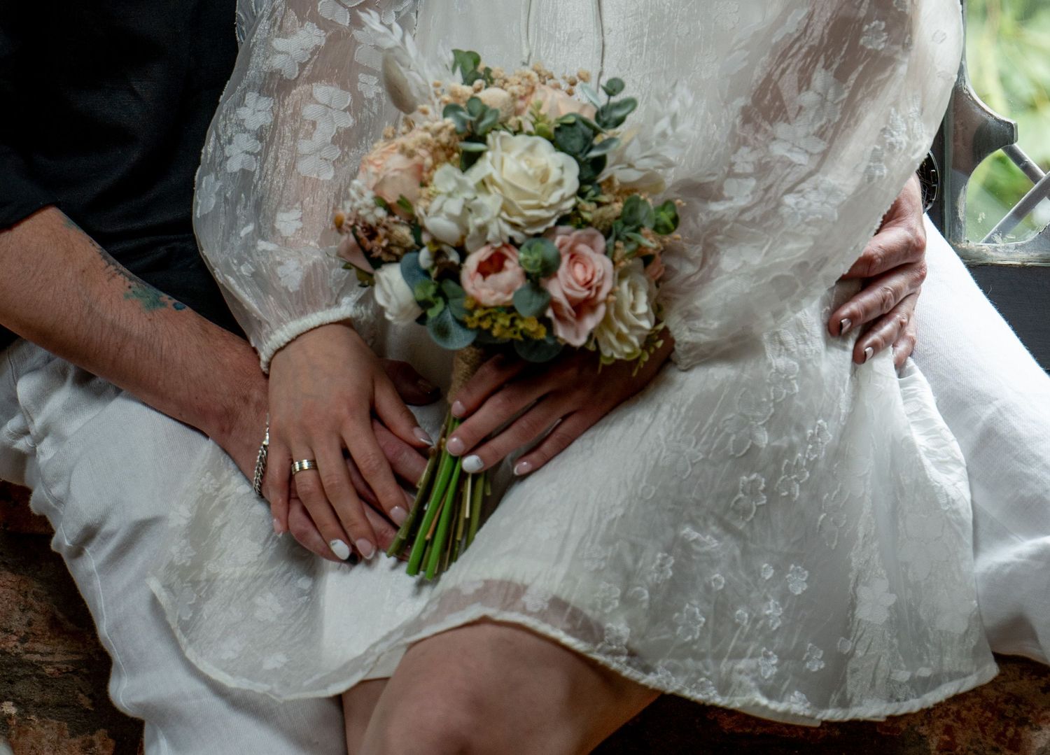 Close-up of a delicate bridal bouquet with peach roses and greenery against a white lace dress.