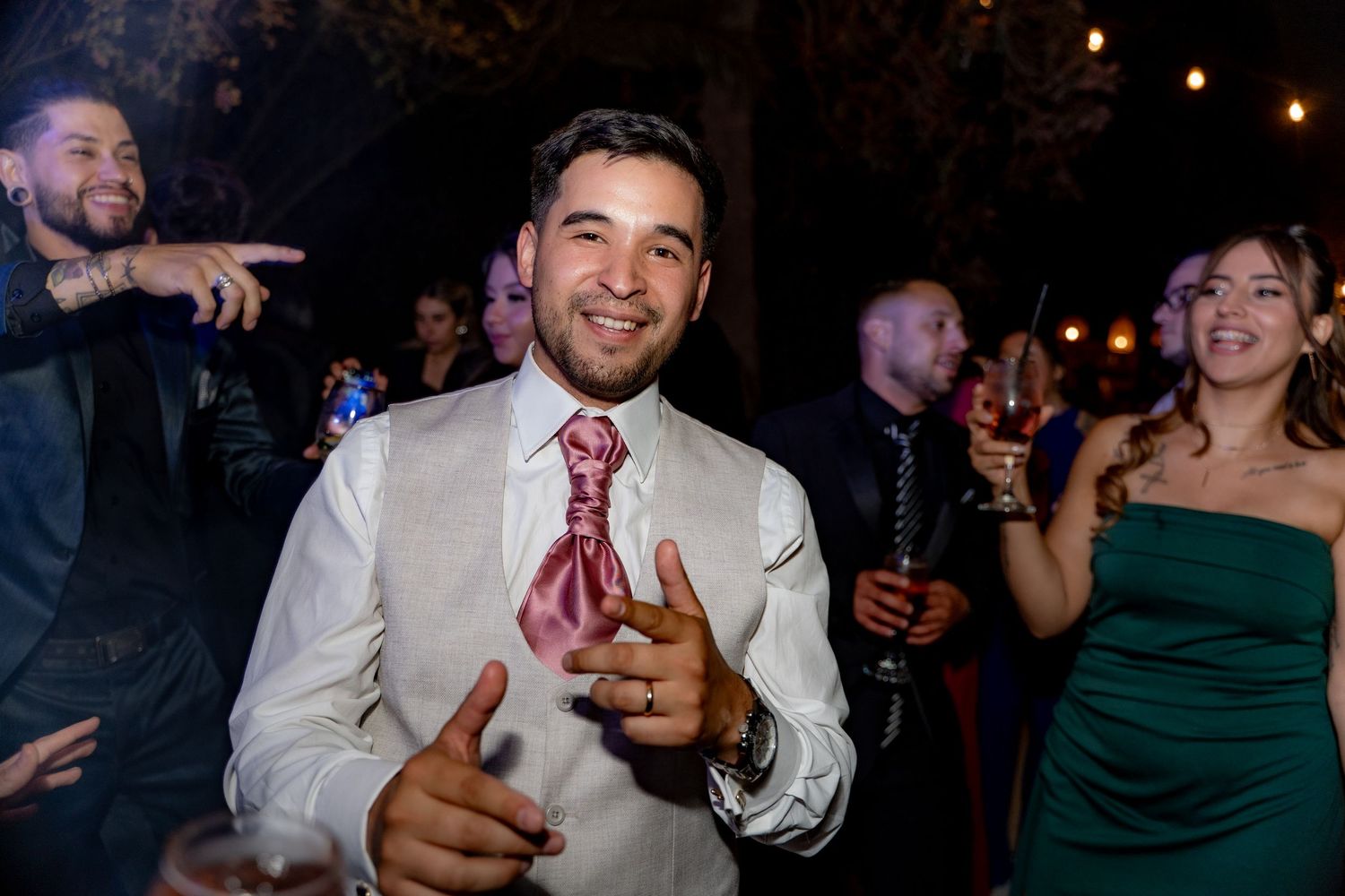 Man in white dress shirt and pink tie smiling at wedding reception party with guests in background.