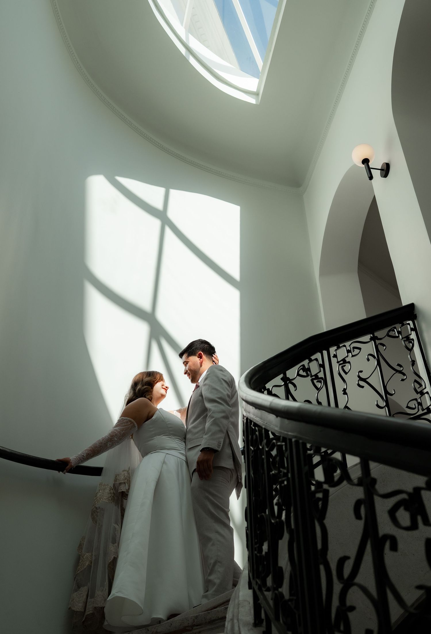 Bride and groom share a romantic moment on an elegant curved staircase with ornate black iron railing and skylight.