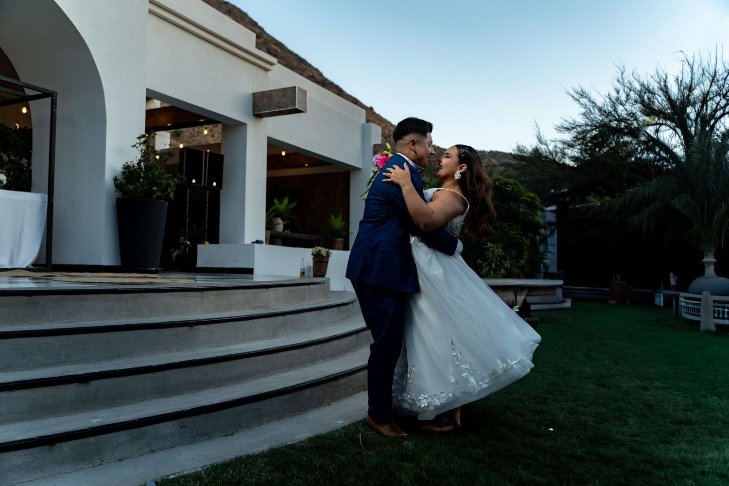Wedding couple embraces on white steps during an outdoor evening celebration.