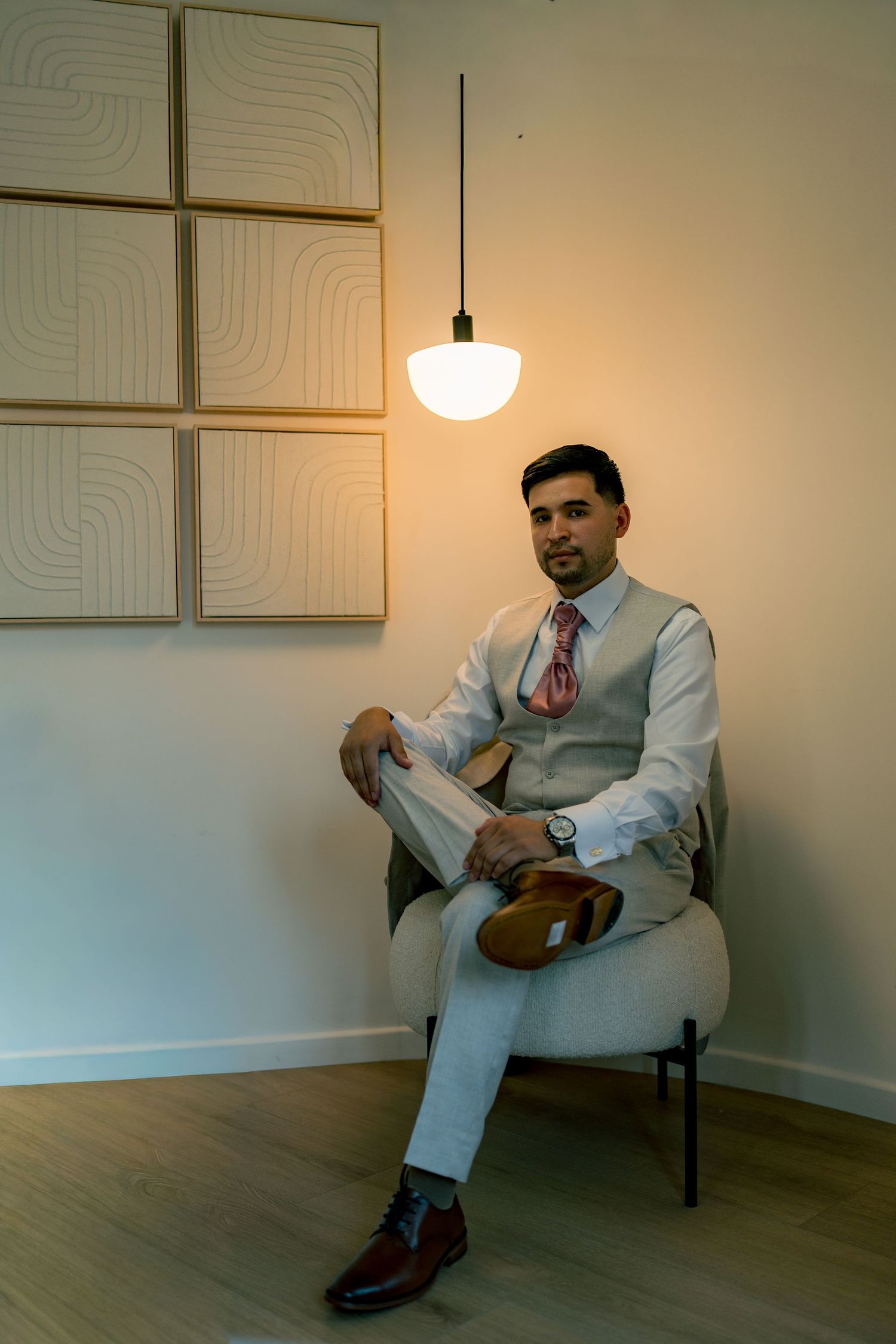 Groom in light gray suit and pink tie seated in modern chair under pendant lamp.