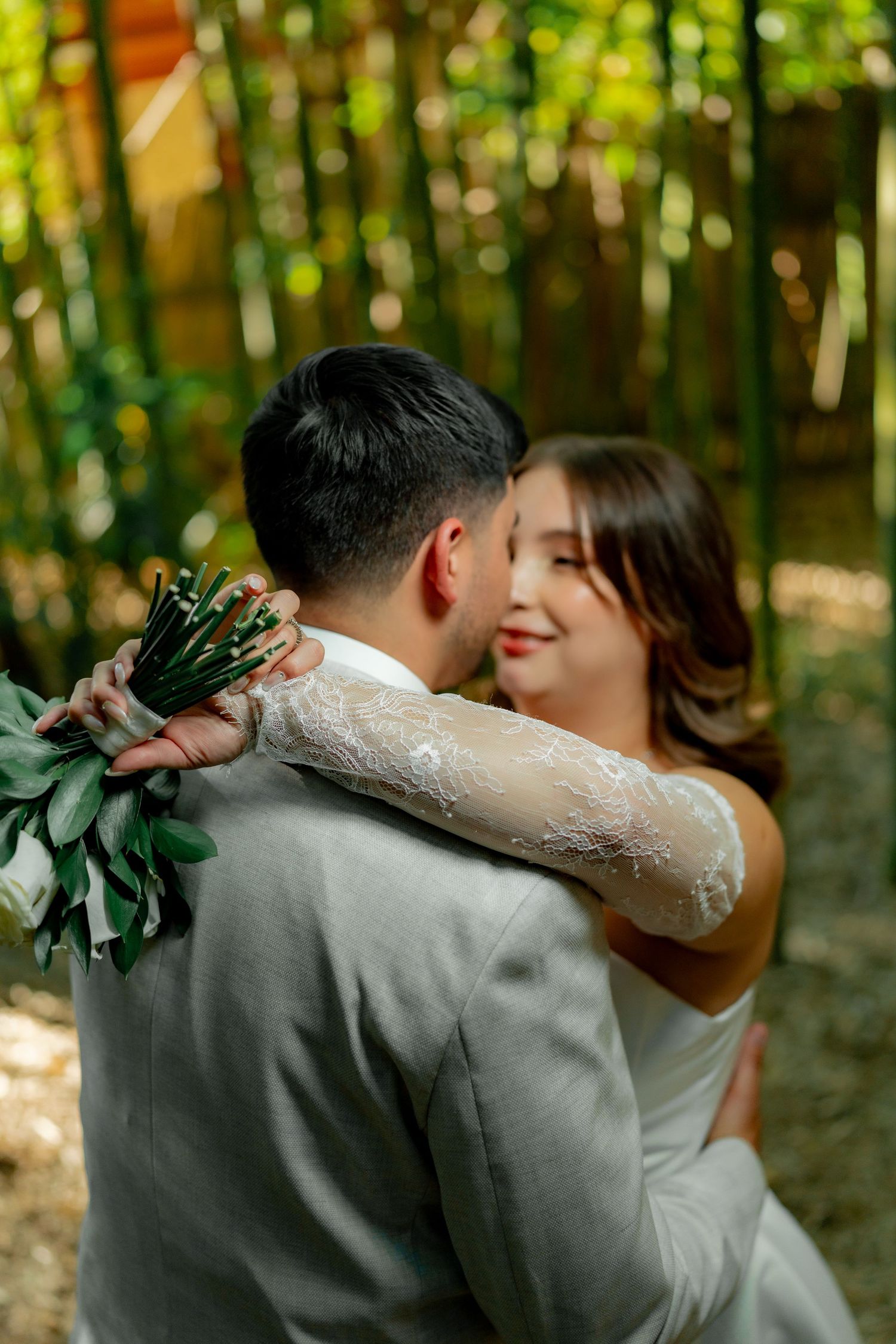 A bride in lace gown embraces groom in gray suit, holding bouquet, in a lush bamboo garden setting.