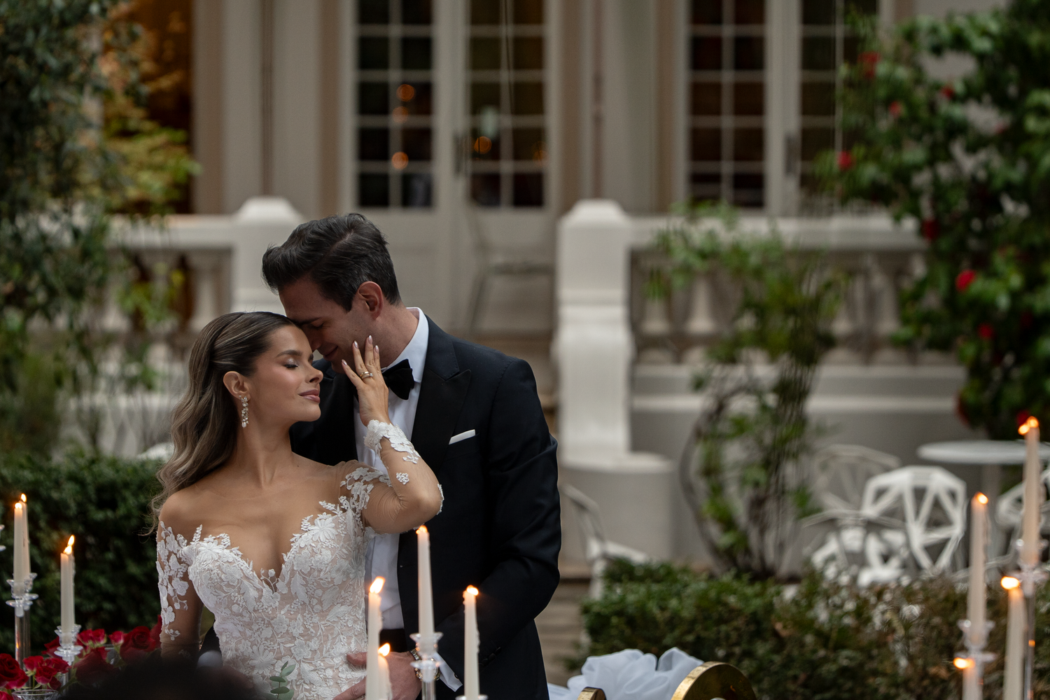 A romantic wedding couple shares an intimate moment in a garden setting with elegant architecture.