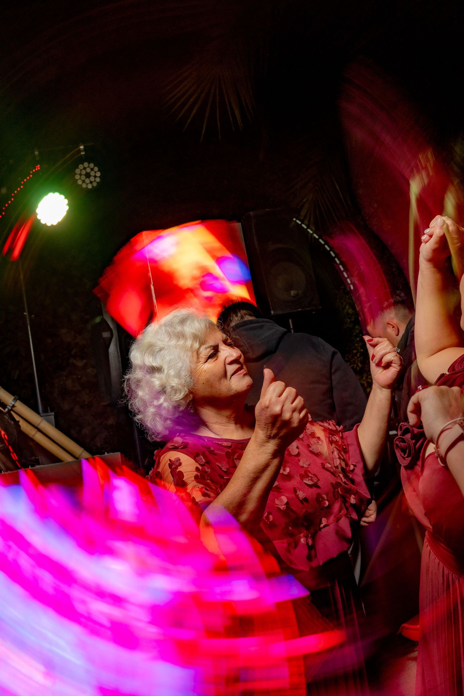 Elderly woman with white curly hair dancing joyfully under colorful lights at a festive wedding party.