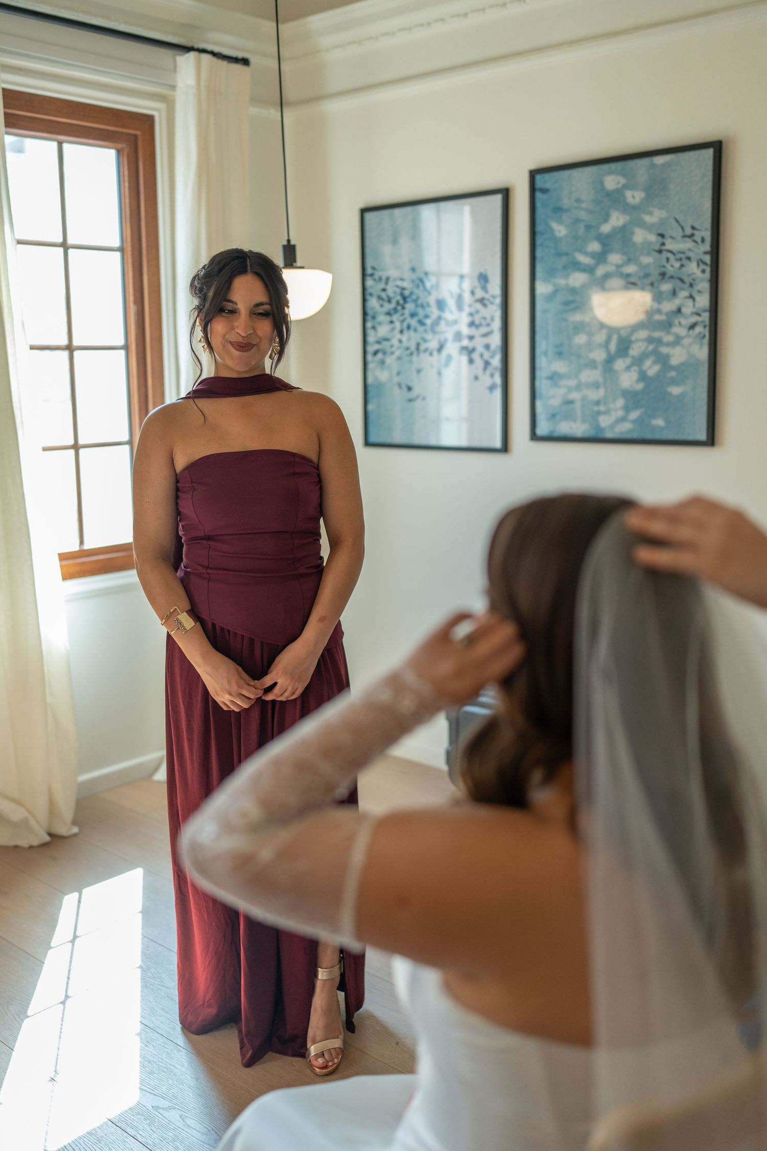 Bridesmaid in burgundy strapless gown watches bride adjust her veil in bright wedding suite.