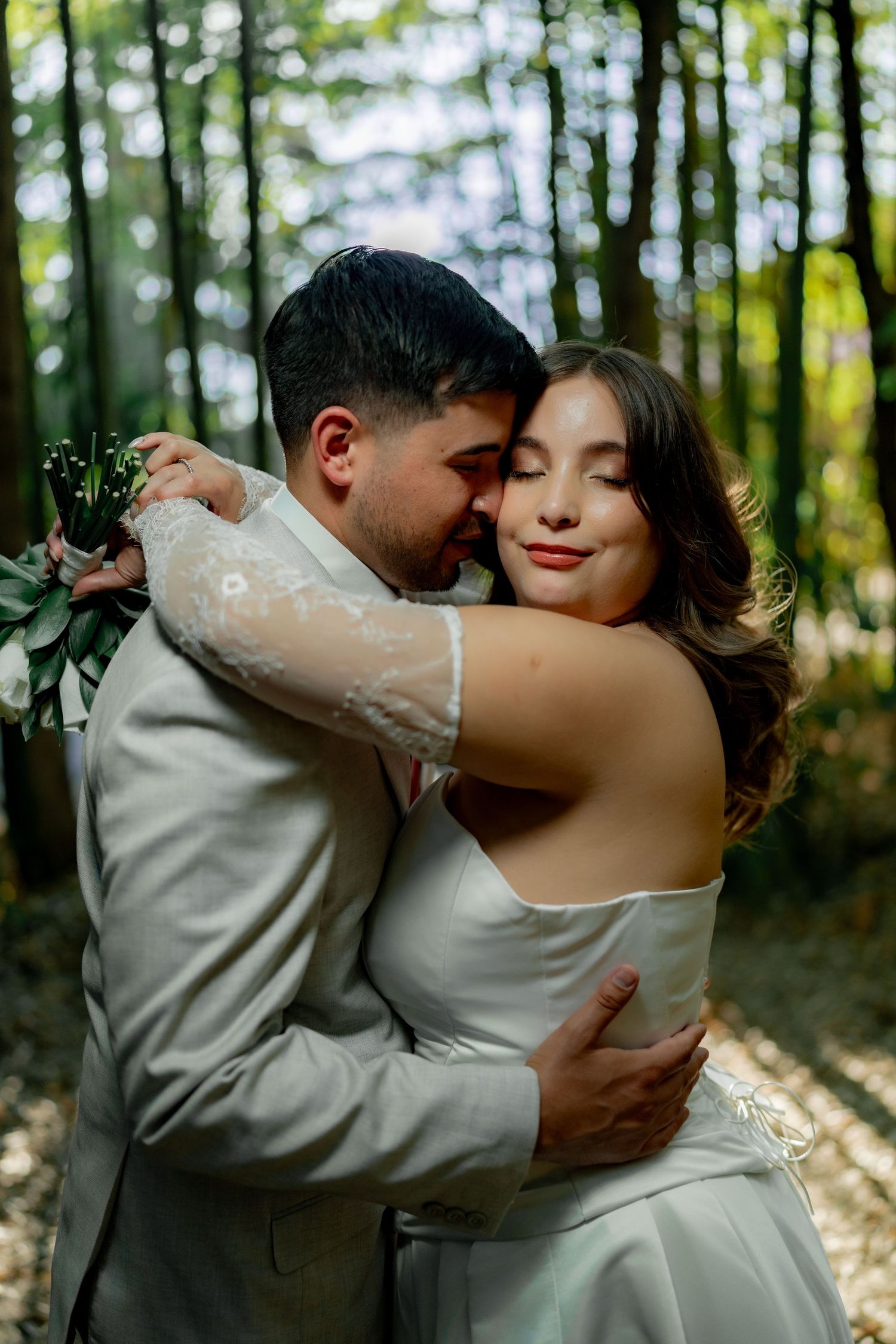 A bride and groom share a romantic embrace in a sunlit forest, with the groom kissing her cheek tenderly.