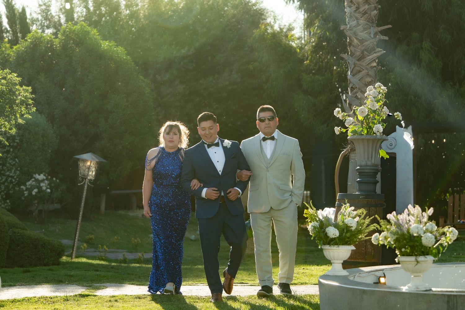 Wedding party members walk together through a sunlit garden path during golden hour.