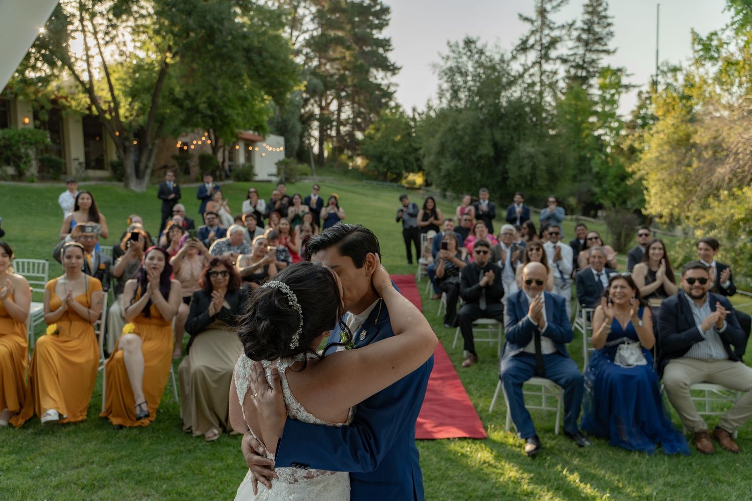 Wedding guests seated outdoors on lawn during ceremony with trees in background.