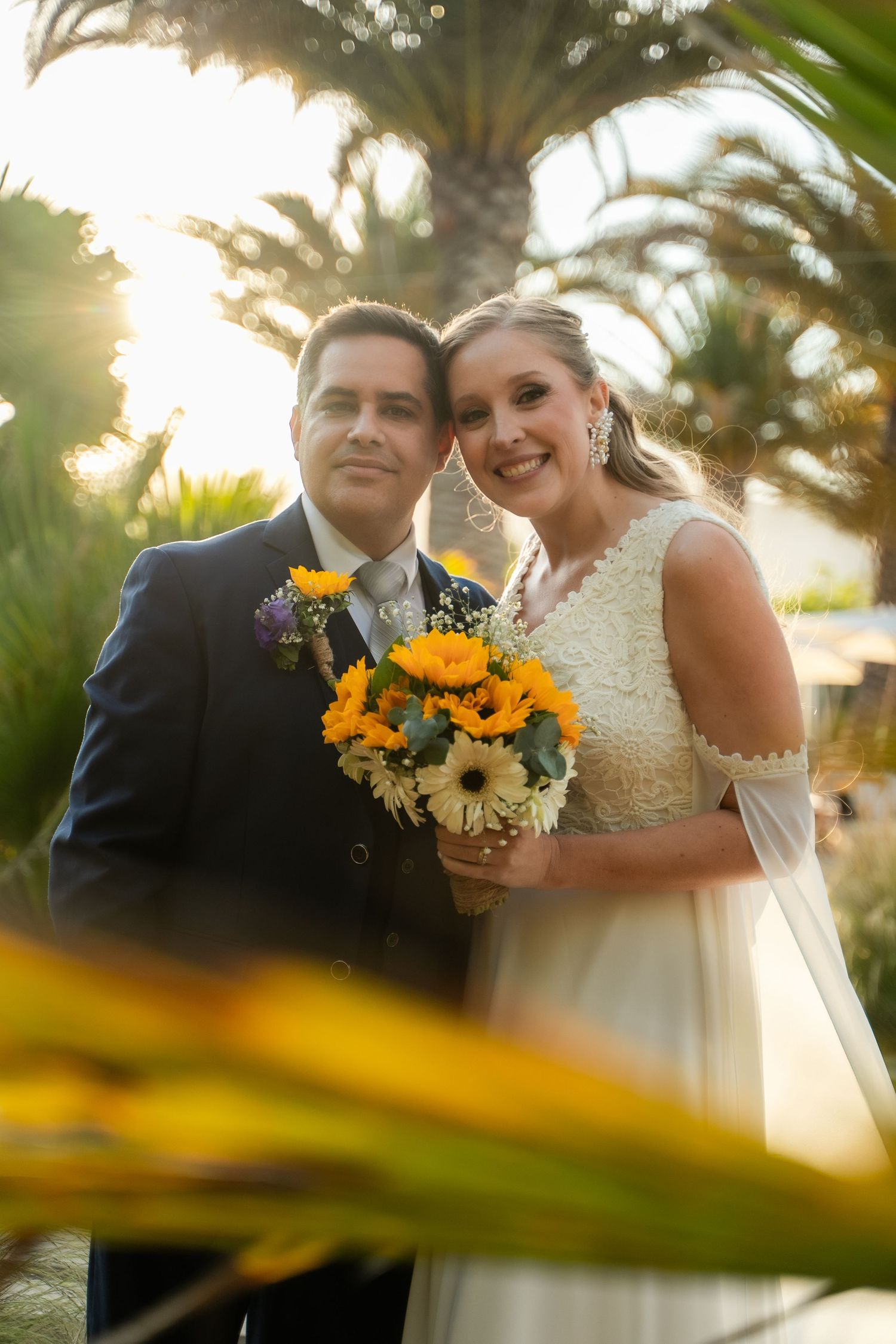 Newlyweds pose with sunflower bouquet against palm trees at sunset.