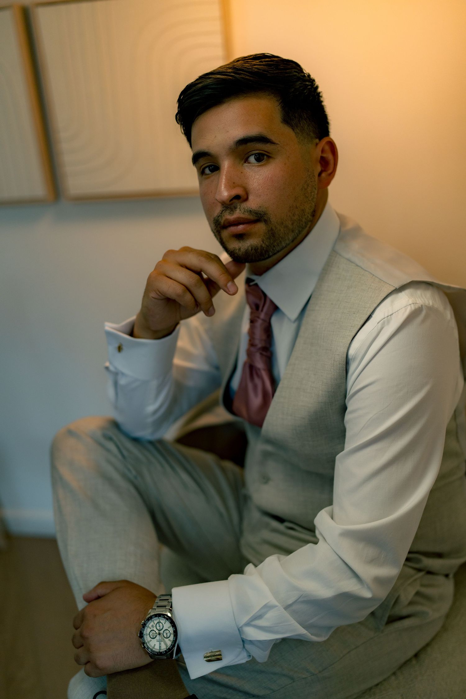 Handsome groom in gray vest and pink tie seated confidently with a luxury watch on his wrist.