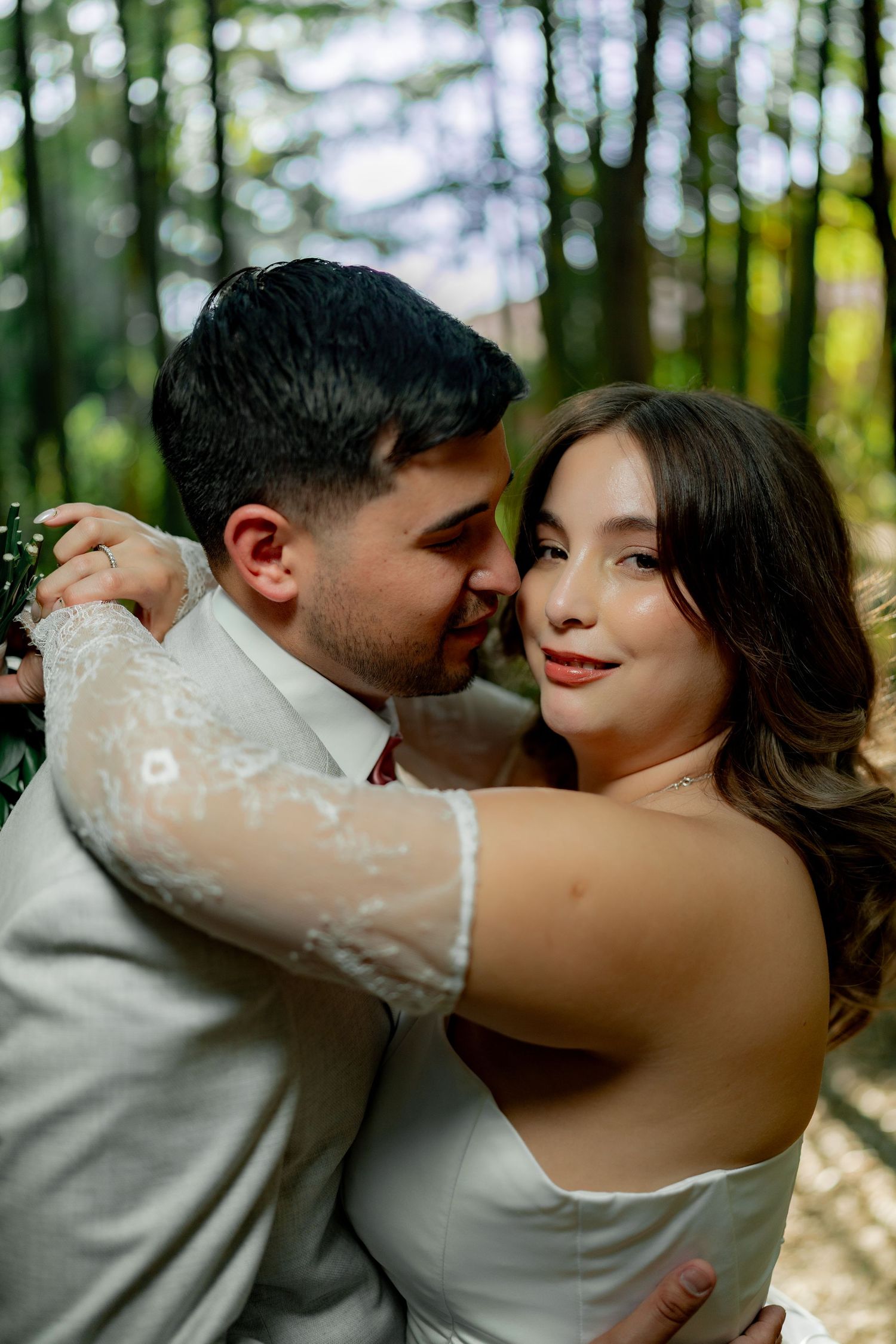 Groom in beige suit embraces bride in white lace gown in sunlit forest setting.