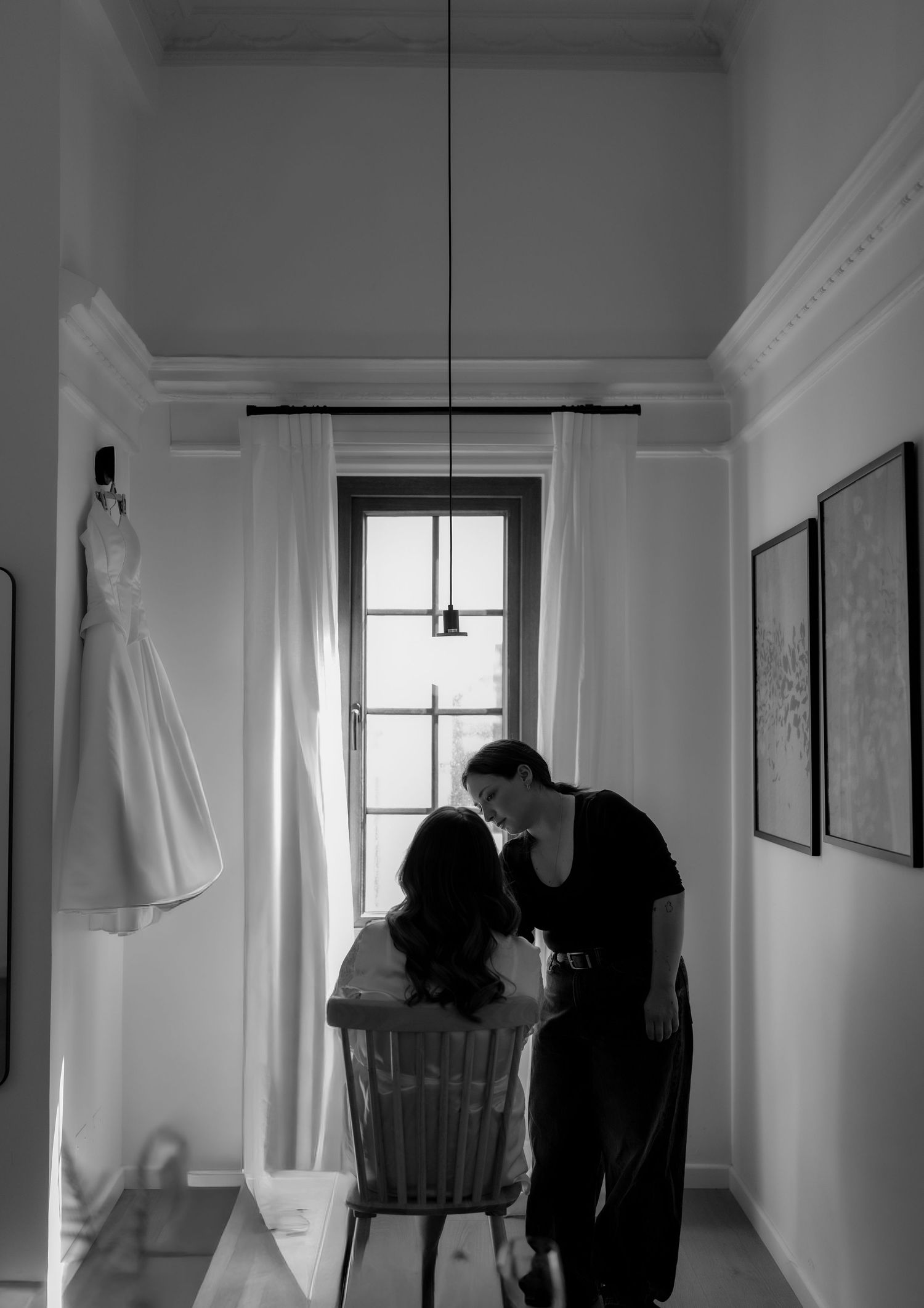 Black and white photo of a couple sharing an intimate moment in a room with a wedding dress hanging nearby.